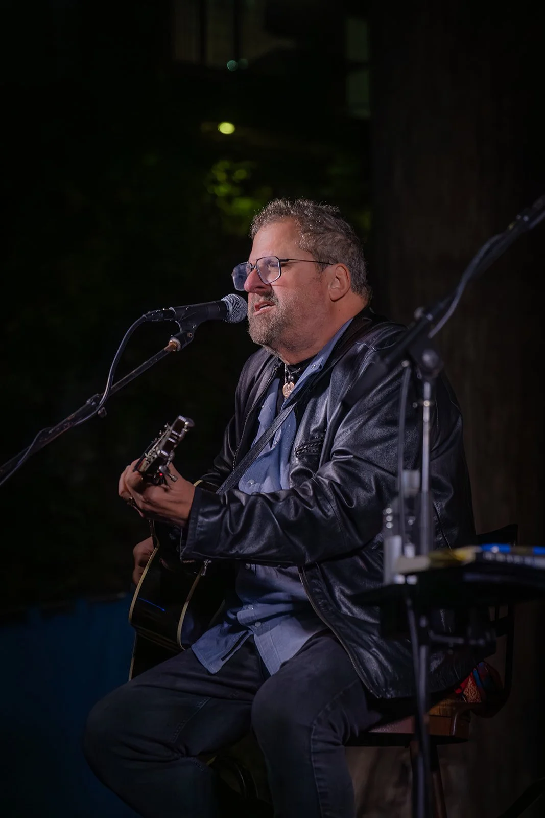 Man with glasses and beard singing into a microphone while playing an acoustic guitar at night.