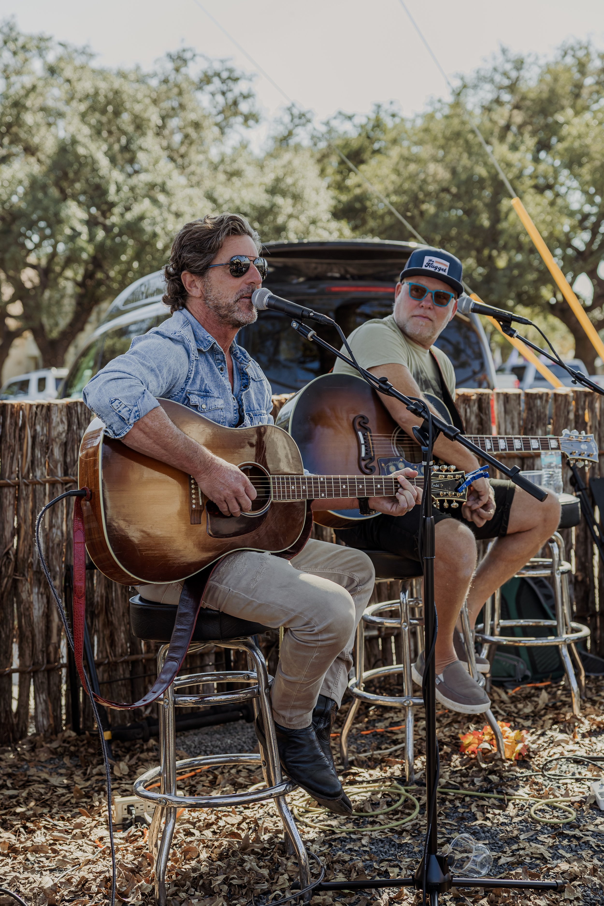 Two men playing acoustic guitars and singing into microphones outdoors, sitting on bar stools in front of a rustic wooden fence with a background of trees.