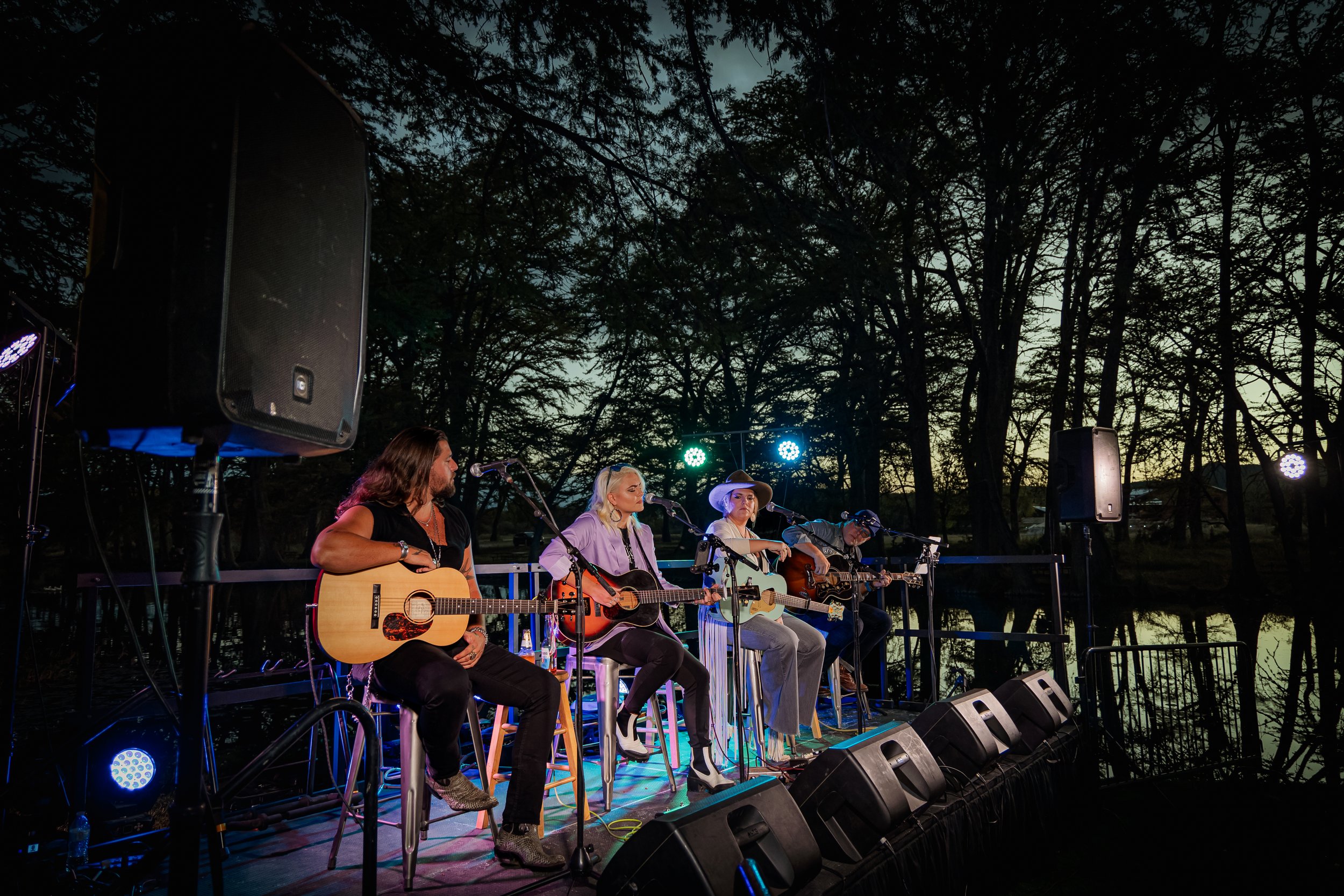 Four musicians playing guitars on an outdoor stage at sunset, surrounded by trees.