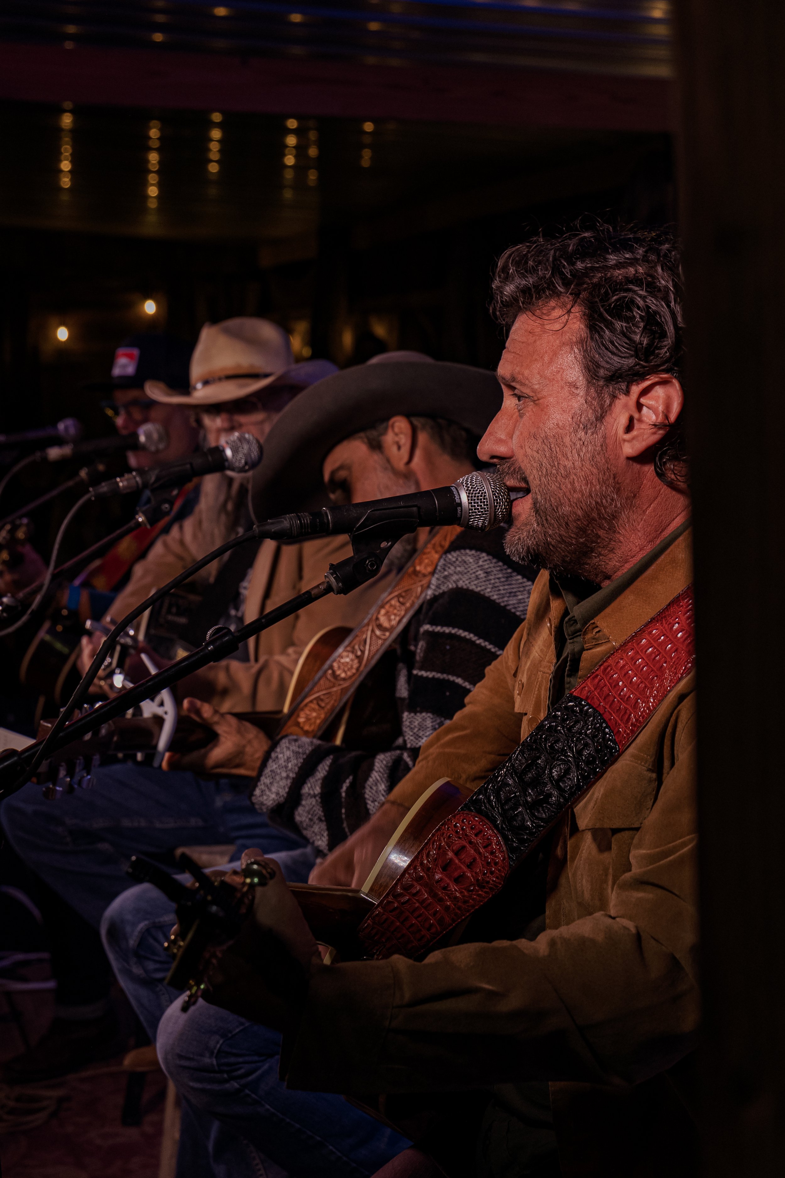 Four men sitting side by side singing into microphones, playing guitars during a live music performance in a dimly lit indoor setting.