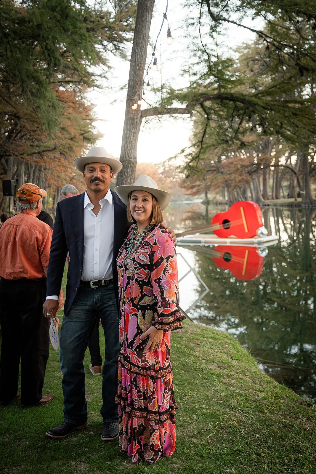 A man and woman in cowboy hats posing by a lake with a red guitar-shaped boat reflected in the water. Other people are in the background near the trees.