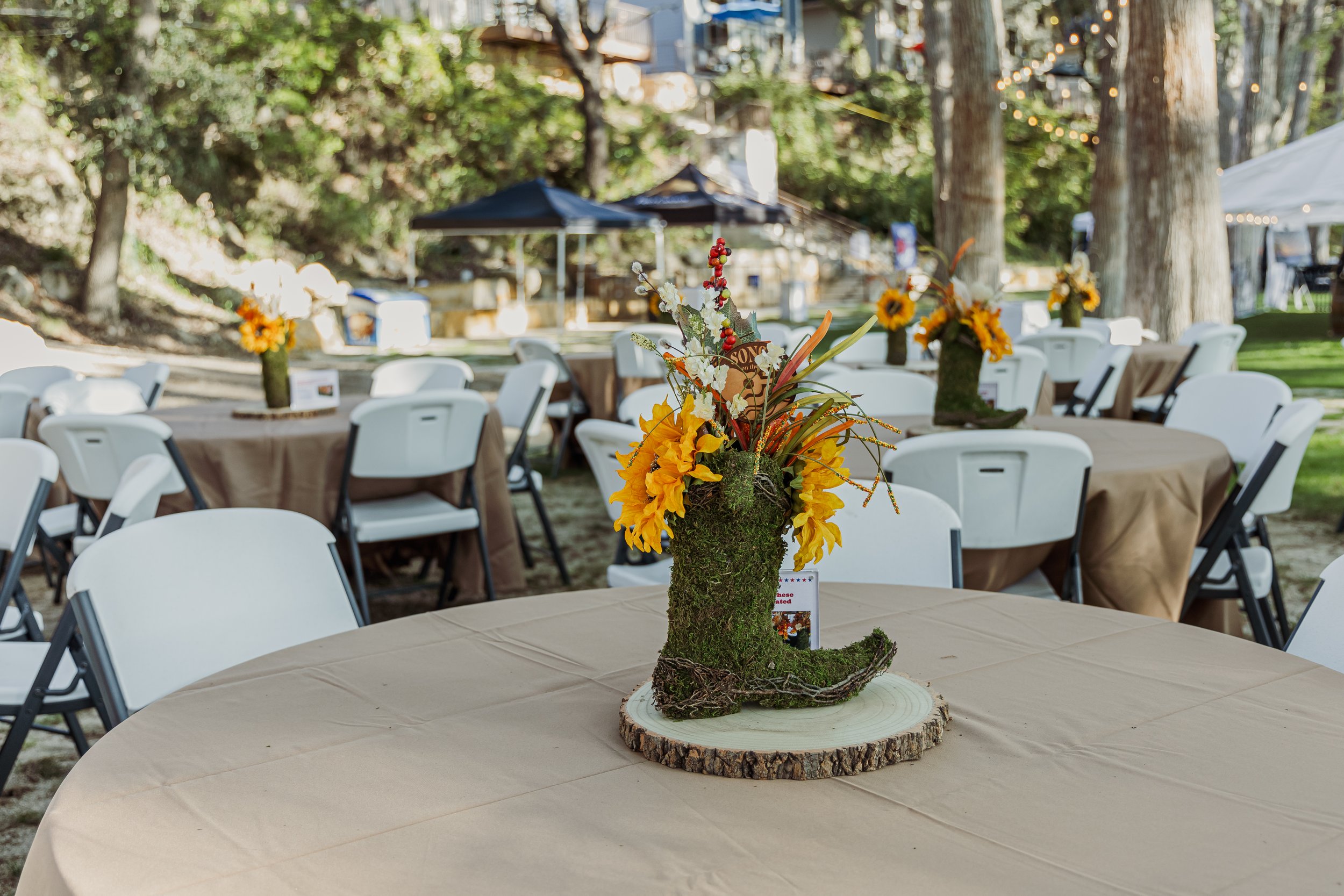 Outdoor event setup with round tables covered in beige tablecloths, decorated with centerpieces resembling yellow boots filled with yellow and white flowers, surrounded by white chairs, with tents and trees in the background.