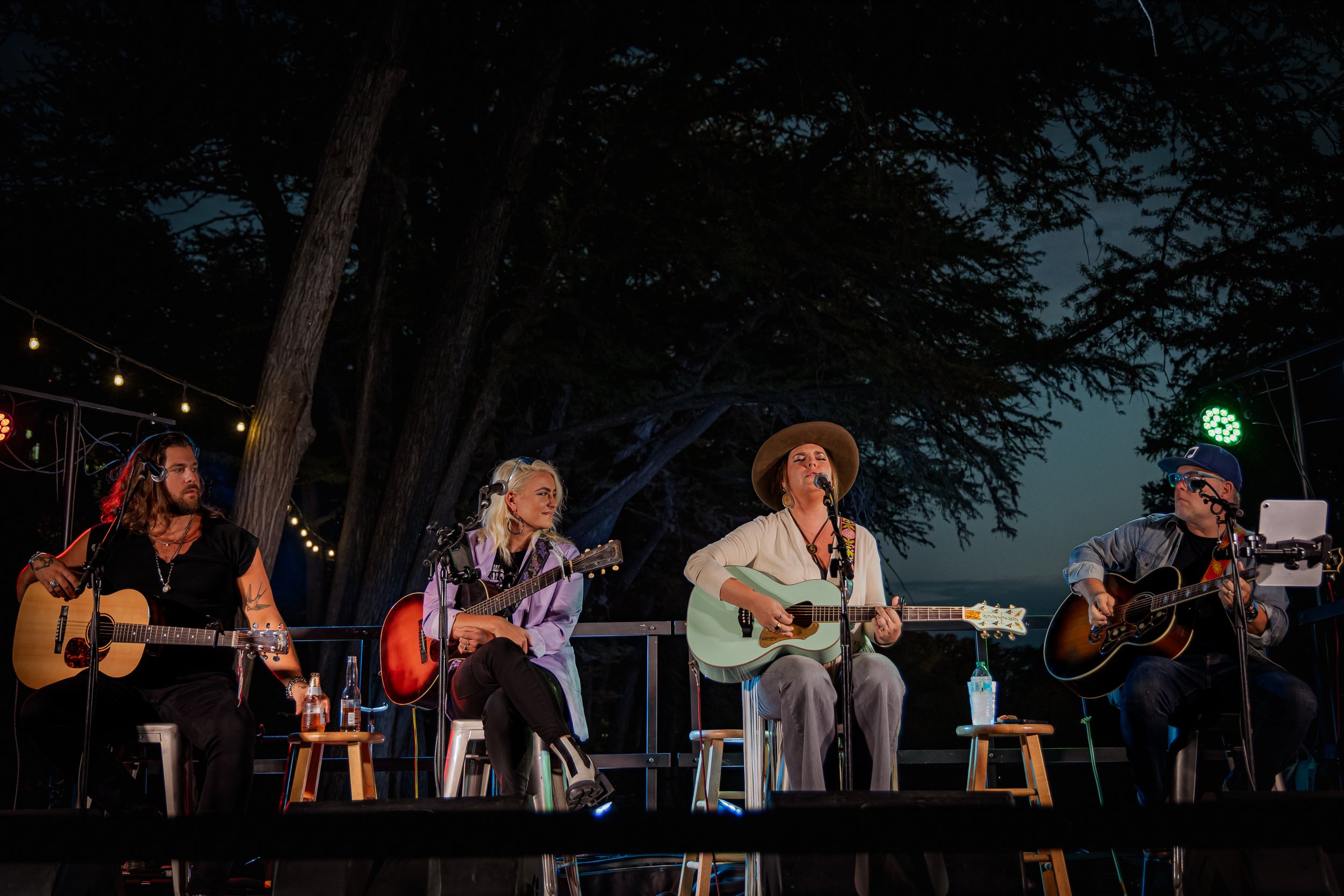 Four people sitting on stage outdoors at night, playing acoustic guitars and singing into microphones, with string lights and large trees in the background.