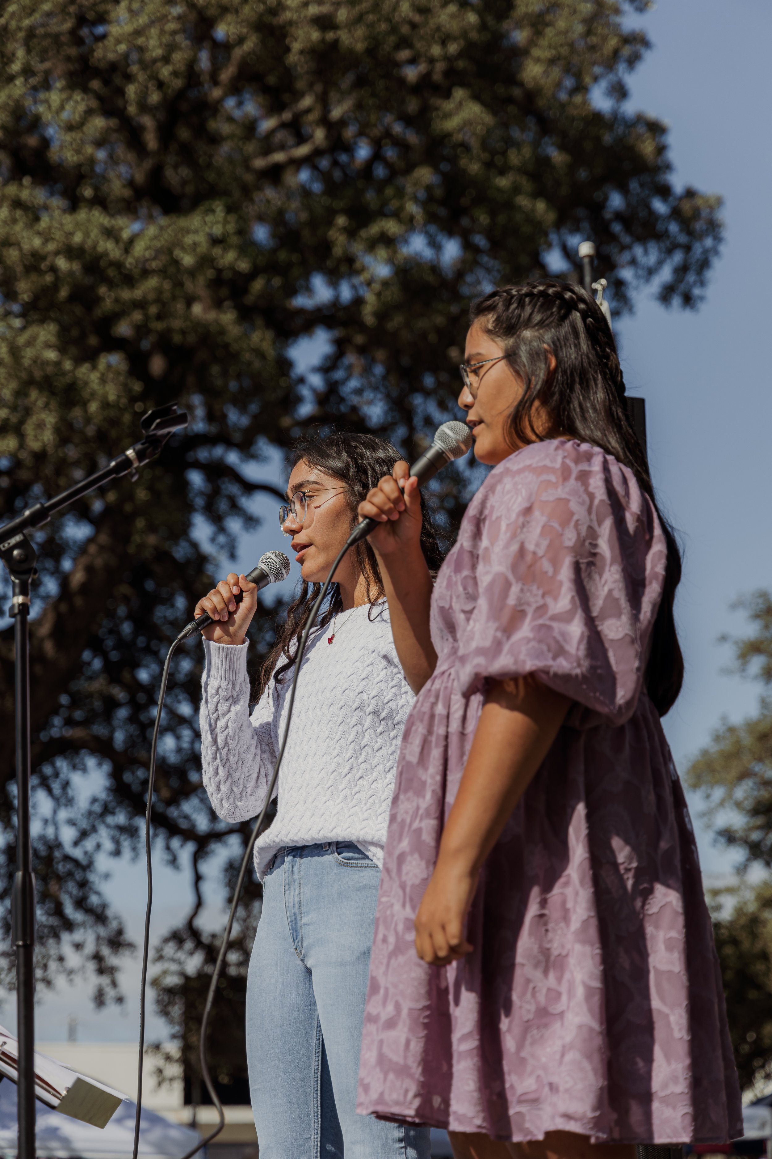 Two women speaking into microphones on an outdoor stage during daylight with a large tree in the background.