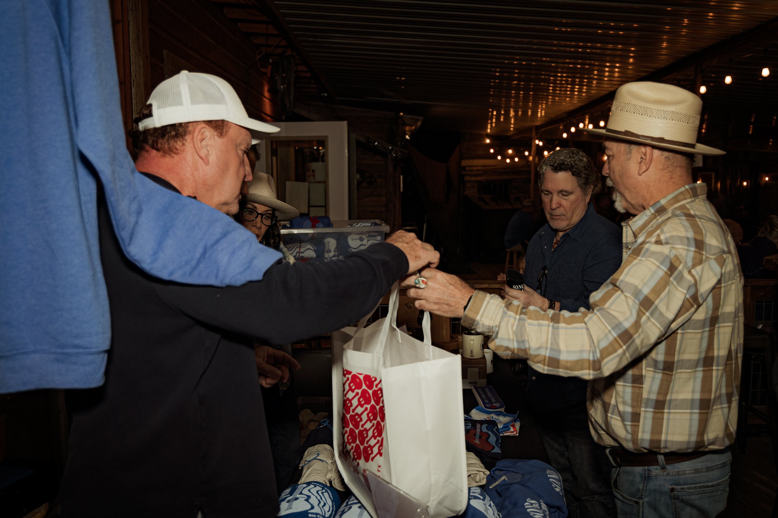 Four people at a store counter exchanging a white shopping bag in a dimly lit, rustic indoor setting