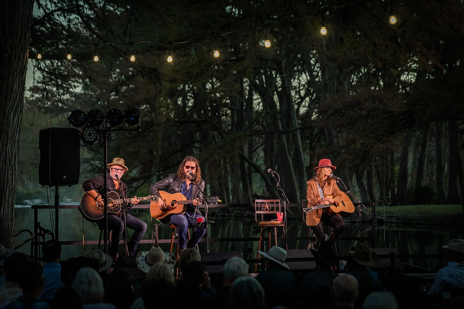 An outdoor musical performance at night with three musicians sitting on chairs on a stage, playing guitars and singing into microphones. The stage is surrounded by trees and string lights, and the audience is seated in front, some wearing hats.