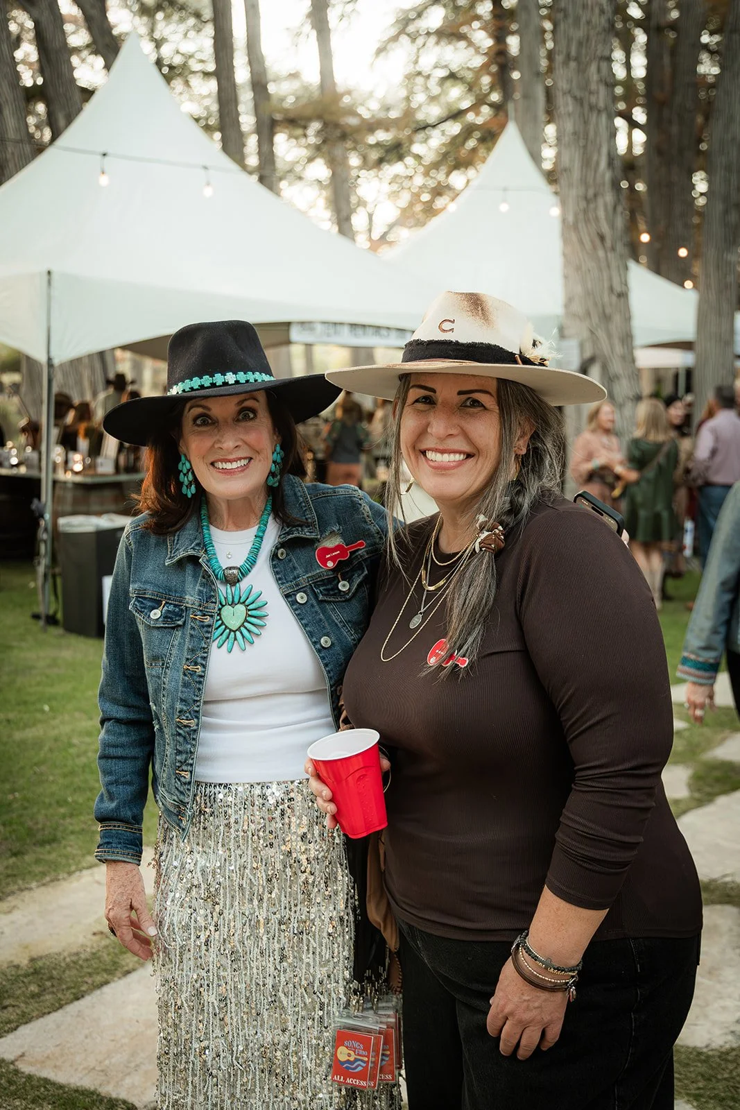 Two women smiling outdoors at a festival or gathering, wearing hats and jewelry, with tents and other people in the background.