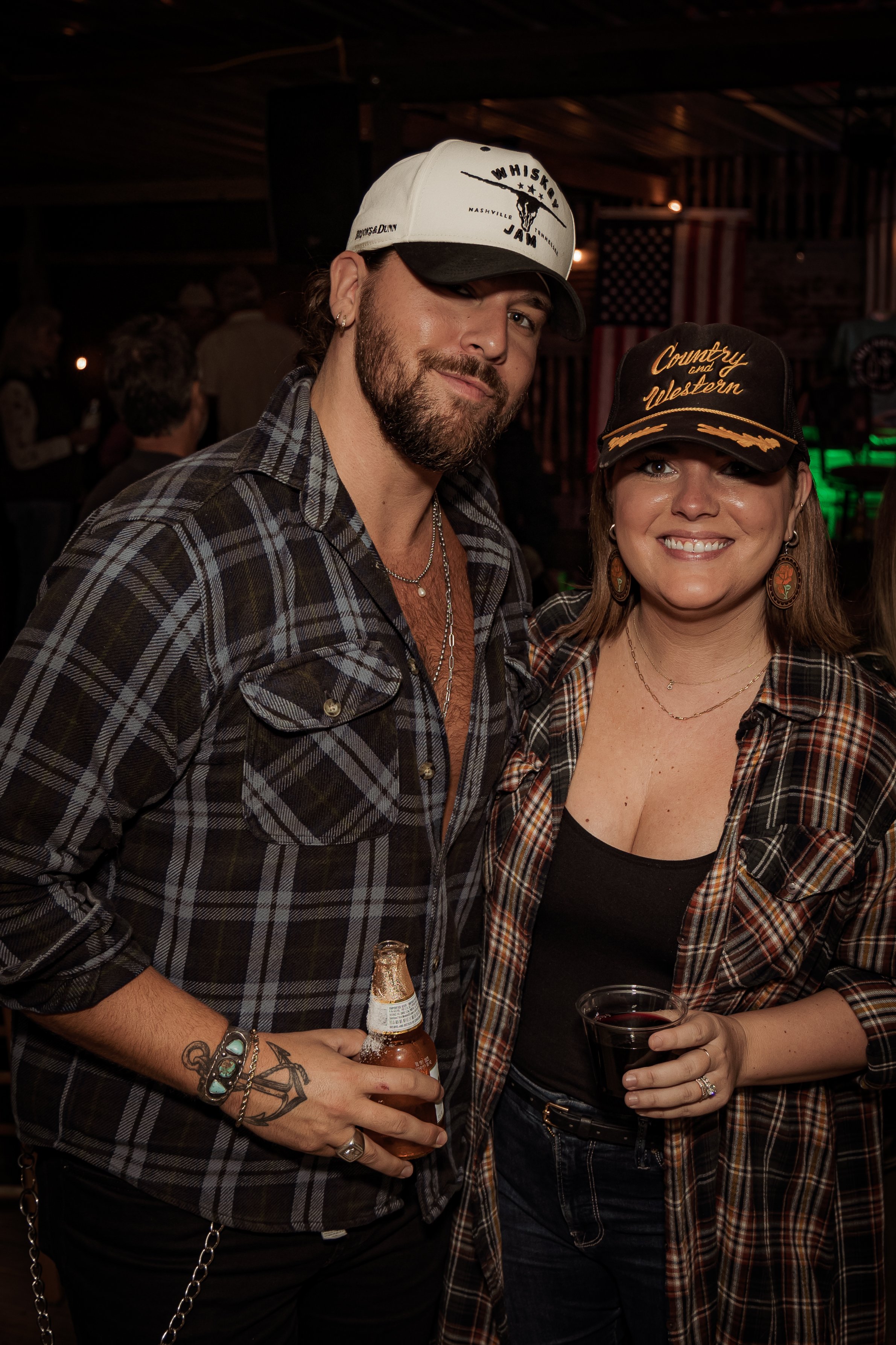 A man and woman in plaid shirts at a bar or concert, smiling with drinks in hand, wearing baseball caps, in a rustic indoor setting with American flag decorations.