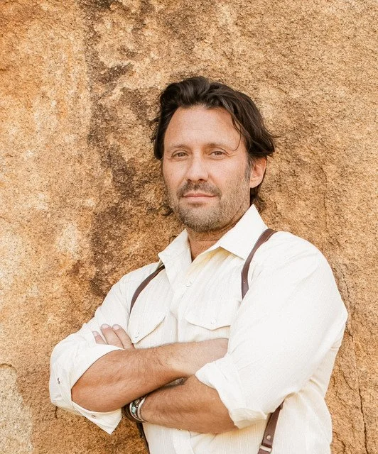 Man with dark hair and beard, wearing a white shirt and suspenders, standing with arms crossed in front of a textured brown wall.