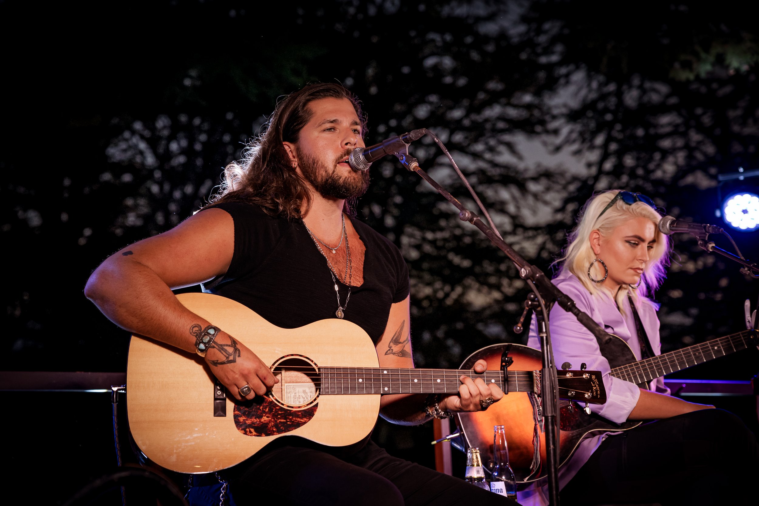 A man with long hair and a beard playing an acoustic guitar and singing into a microphone alongside a woman with blonde hair, wearing headphones and playing an acoustic guitar during a live outdoor performance at night.
