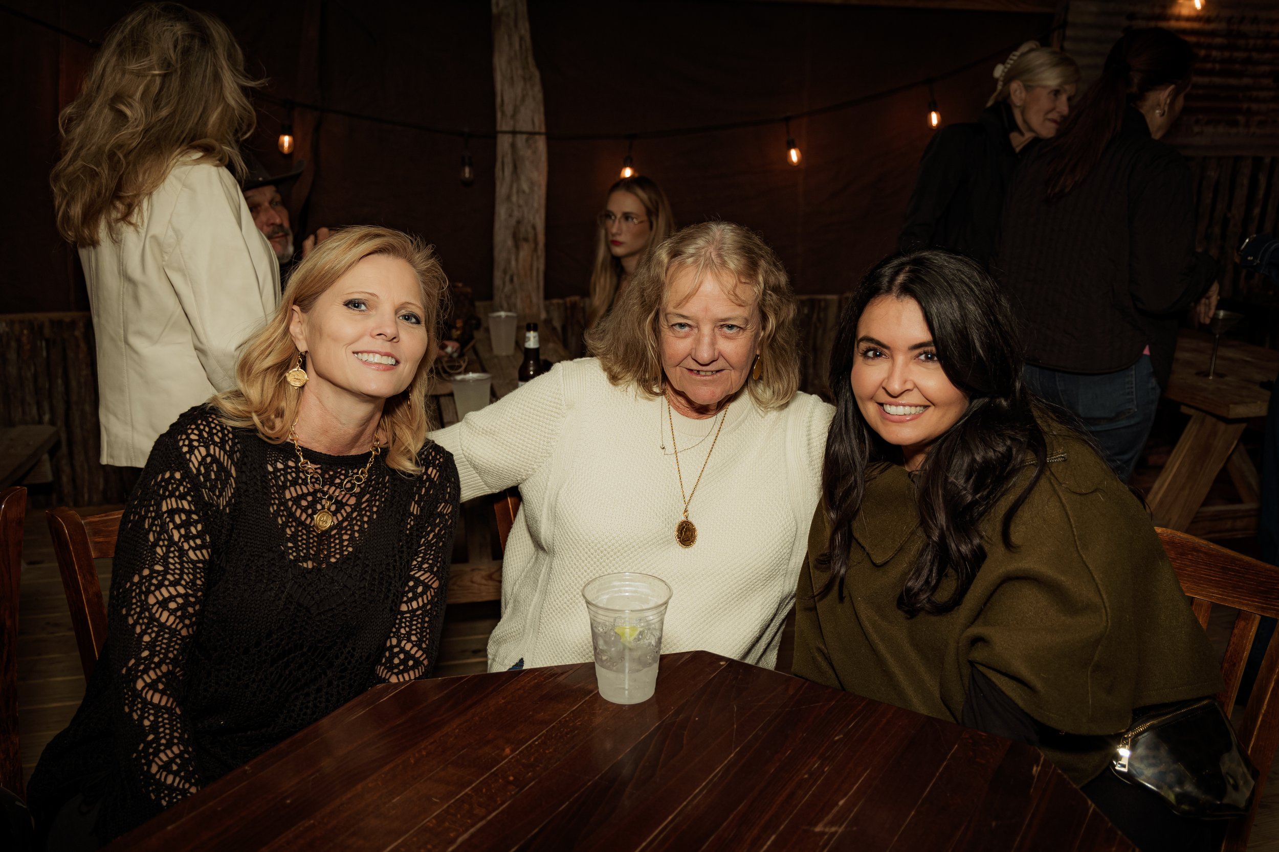 Three women sitting at a wooden table in a dimly lit rustic setting, smiling at the camera, with a plastic cup of water on the table.
