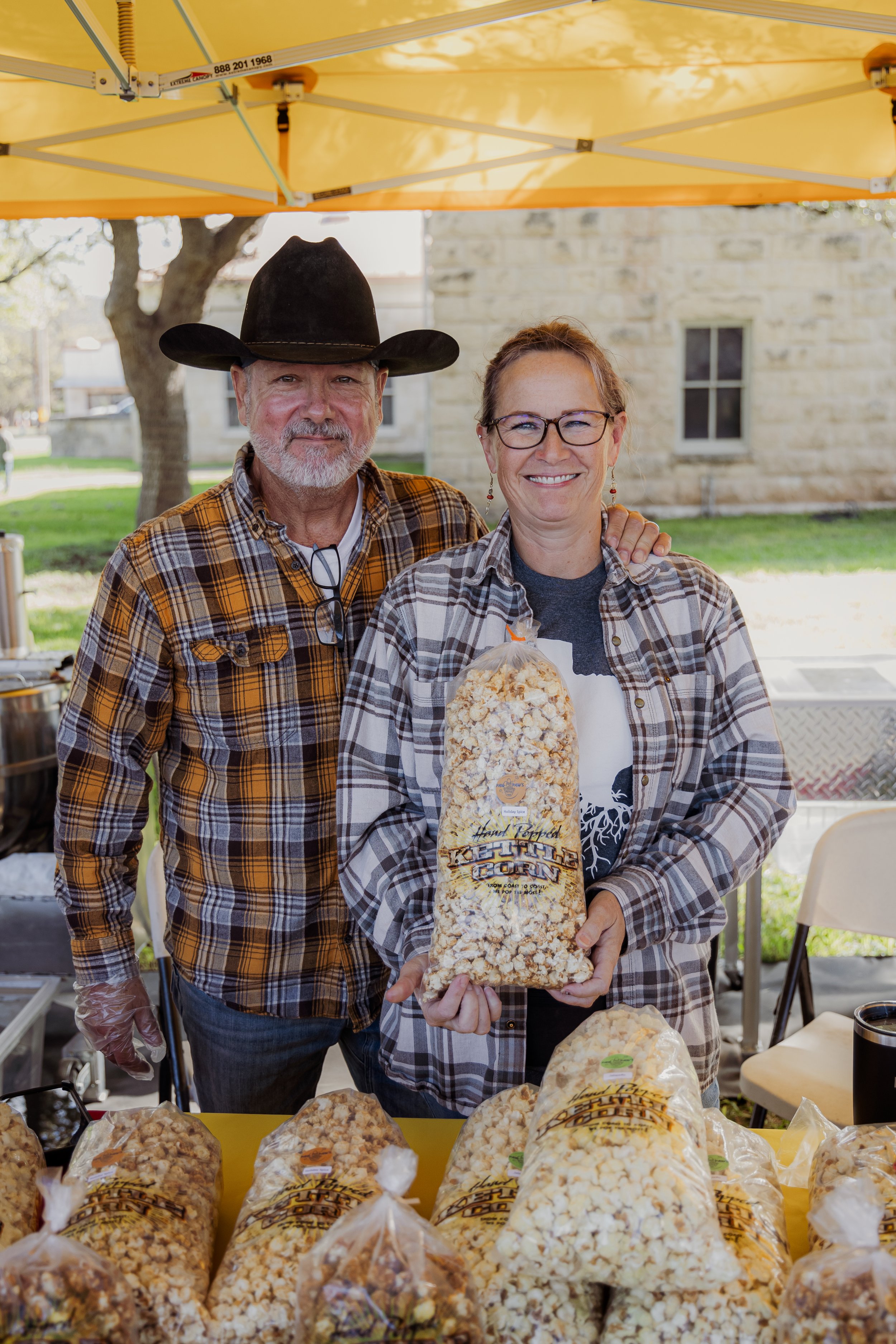 A man and woman at an outdoor popcorn booth, holding a bag of kettle corn, with additional bags of popcorn on the table.