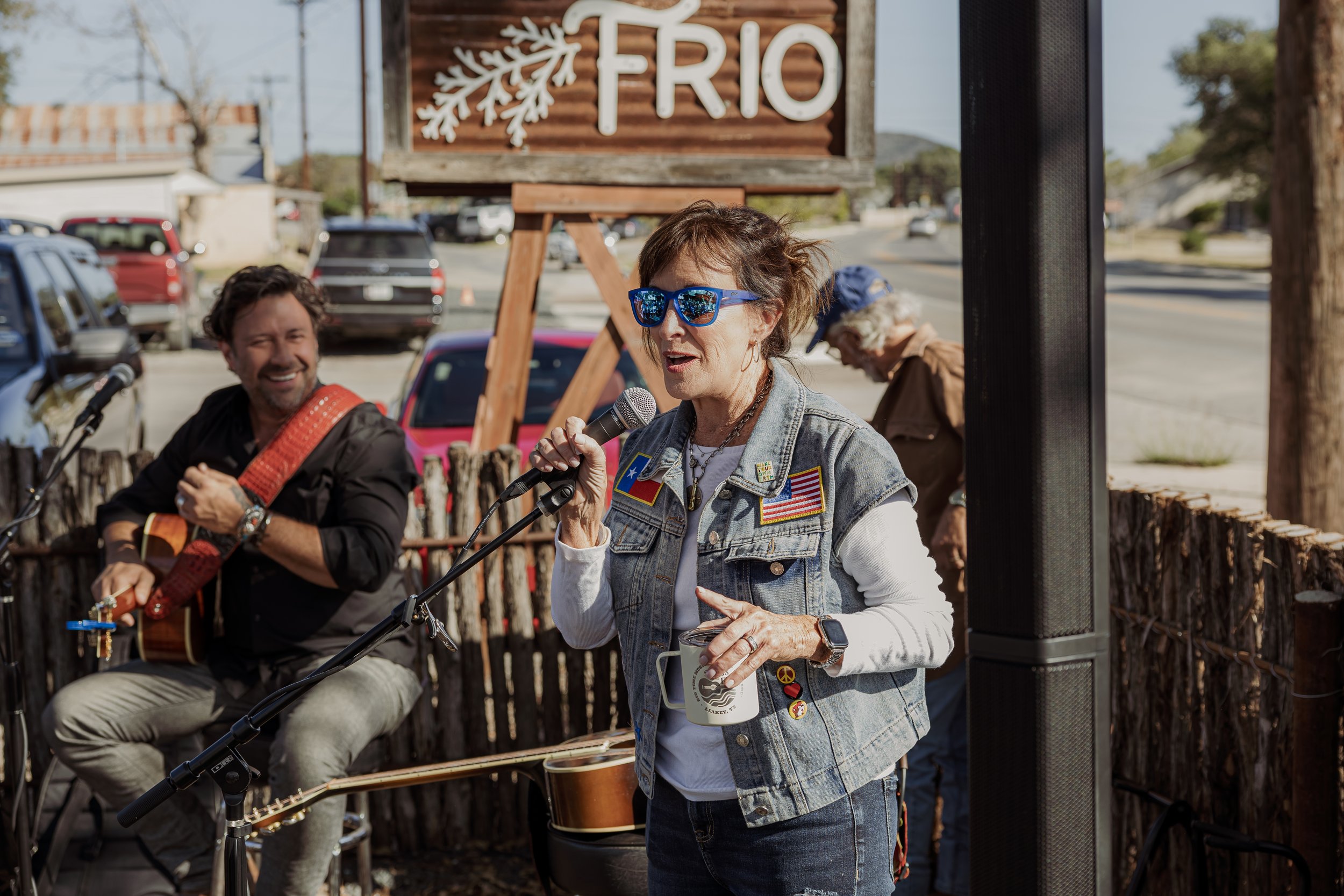 An elderly woman with blue sunglasses singing into a microphone at an outdoor event, with a man playing guitar in the background, in front of a wooden sign that says "FRIO".