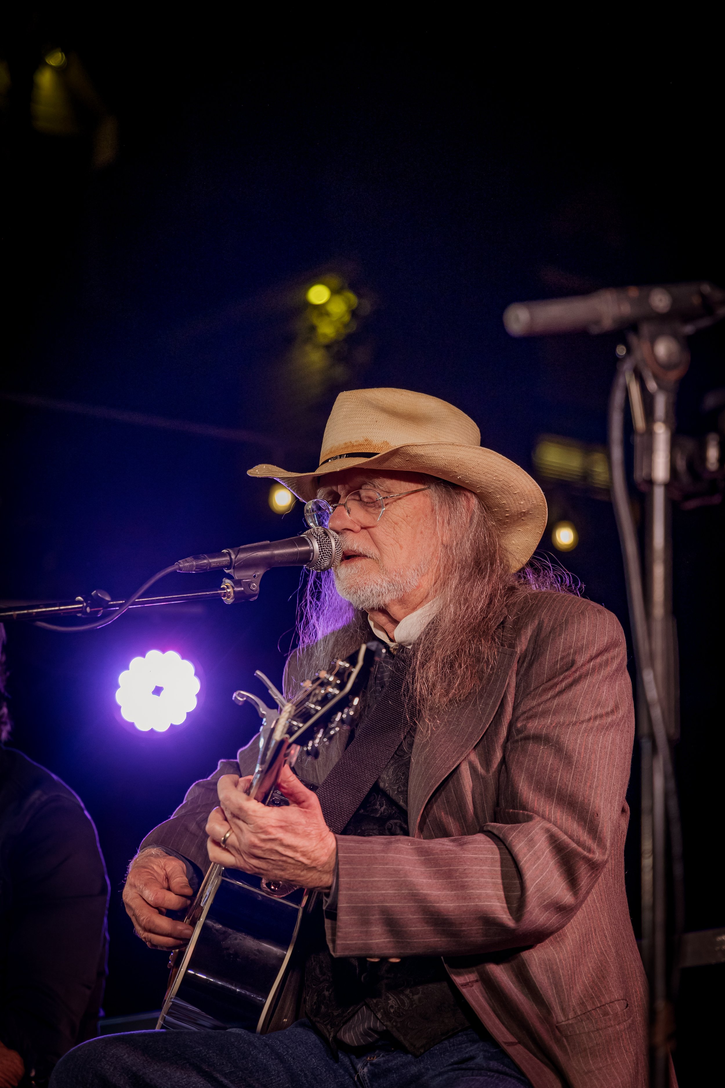 A man with long gray hair and a beard, wearing a beige cowboy hat, glasses, and a brown pinstripe suit, playing an acoustic guitar and singing into a microphone on a stage with purple and yellow lights in the background.