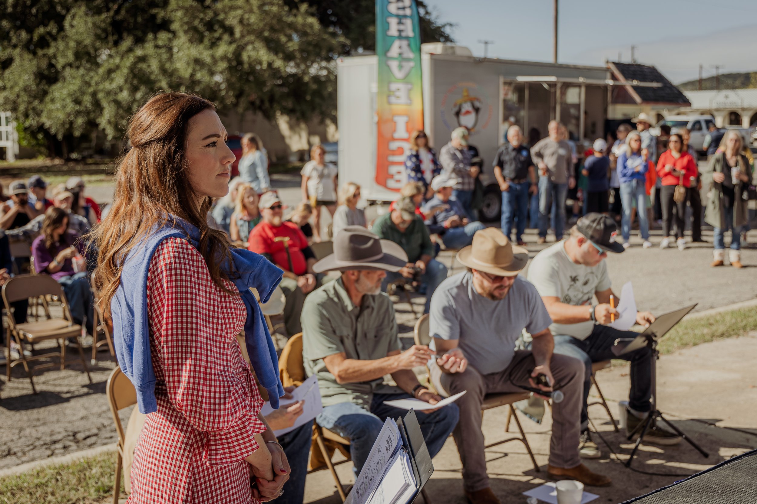 People attending an outdoor event, with a woman in the foreground in a red checkered dress and others sitting and standing, some looking at screens or papers, in front of a food truck and a crowd on a sunny day.