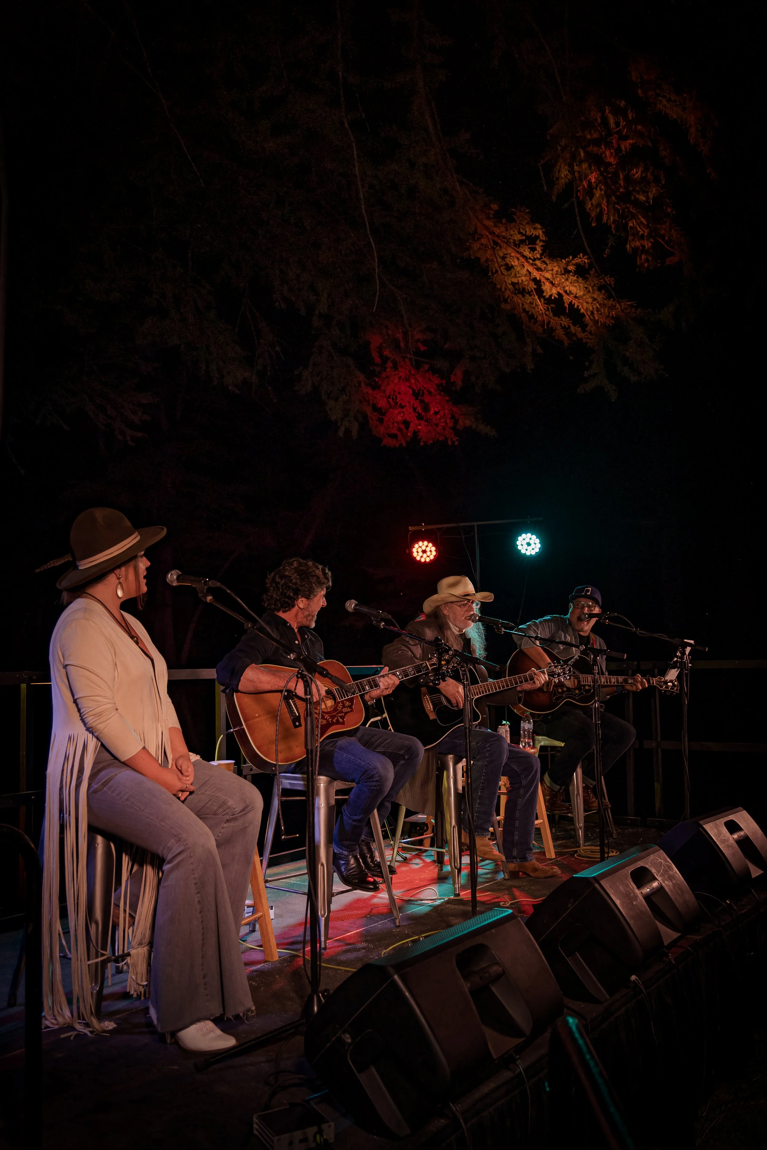 Four musicians performing on an outdoor stage at night, three playing guitars and one singing, with colorful stage lights and a large tree in the background.