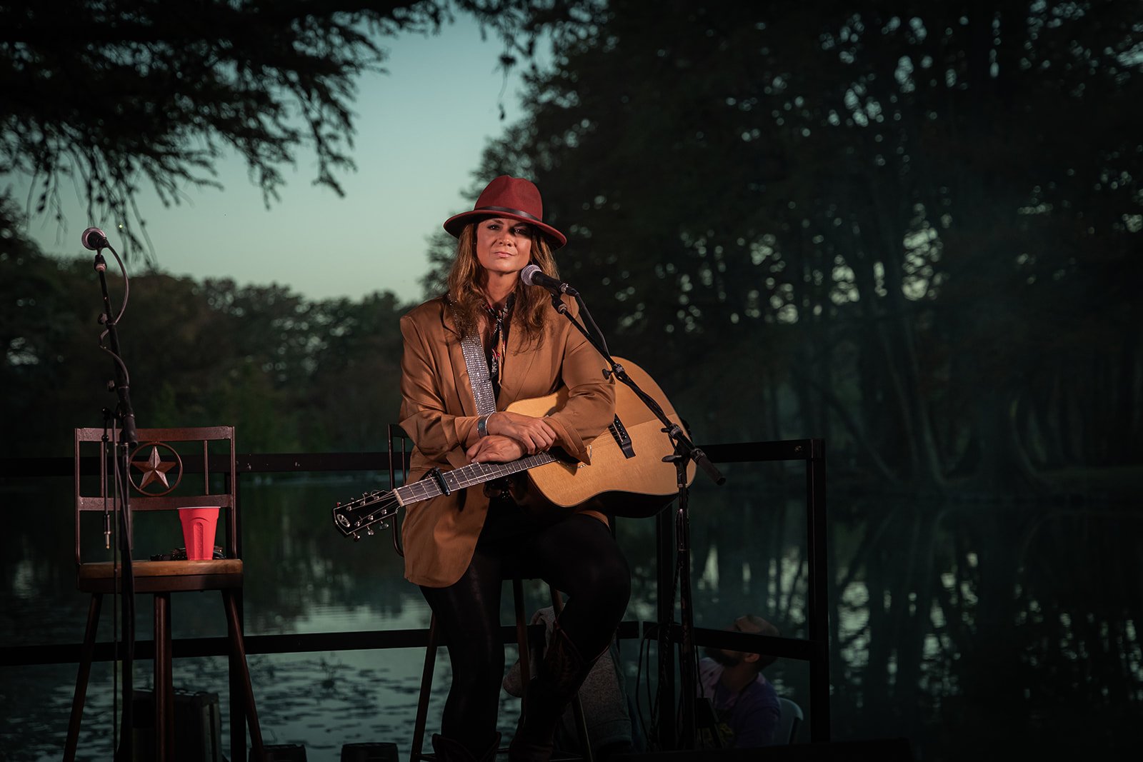 A woman sitting outdoors with a guitar, wearing a brown jacket and a red hat, in front of a scenic water and forest background at dusk.