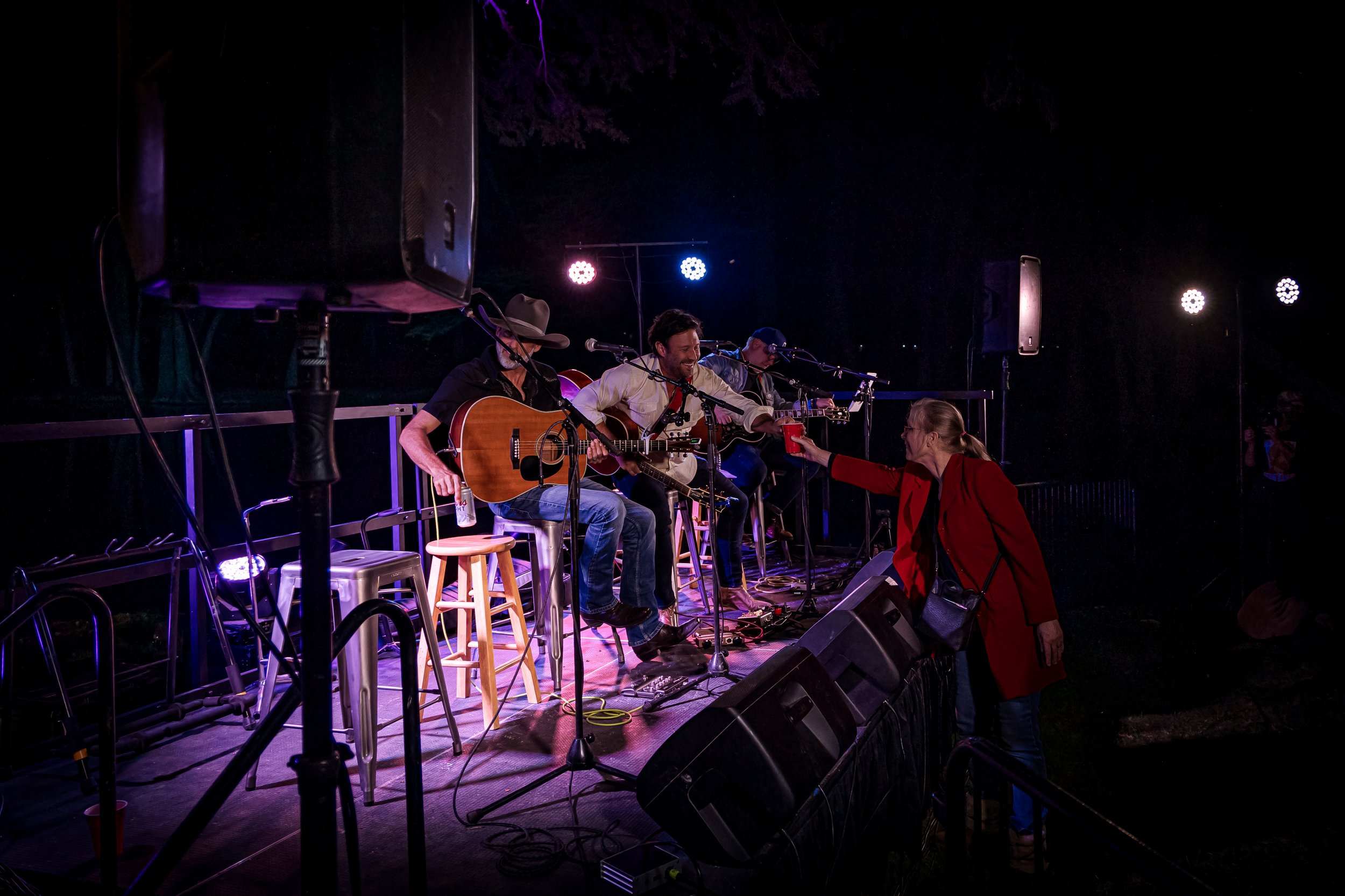 Musicians performing on stage at a nighttime outdoor concert, with one woman in a red coat approaching the stage to hand a drink to a musician.