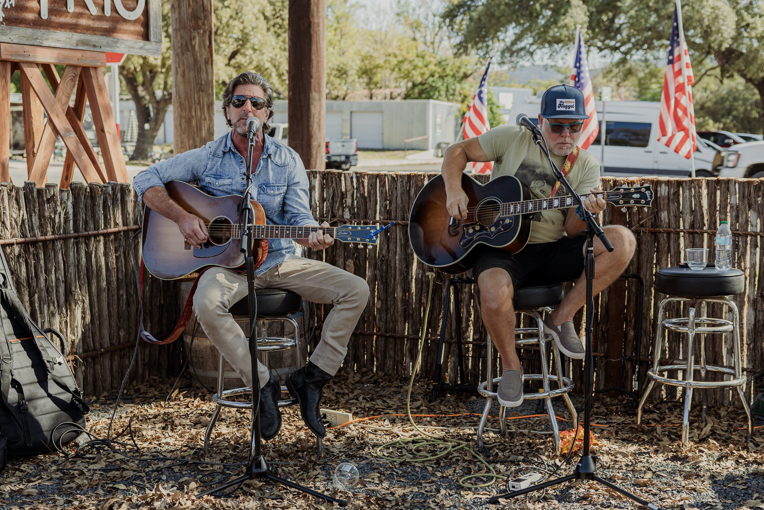 Two men playing guitars on a small outdoor stage with American flags in the background.
