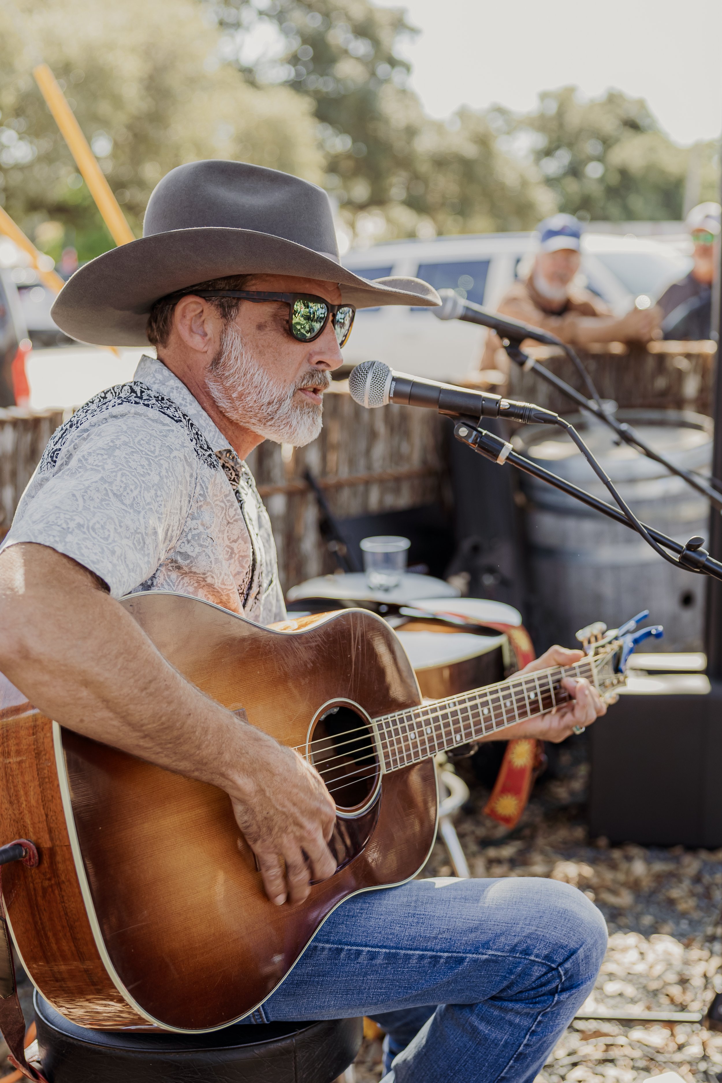 A man wearing a wide-brimmed hat, sunglasses, and a patterned shirt is playing an acoustic guitar and singing into a microphone at an outdoor event. There are people and trees in the background.