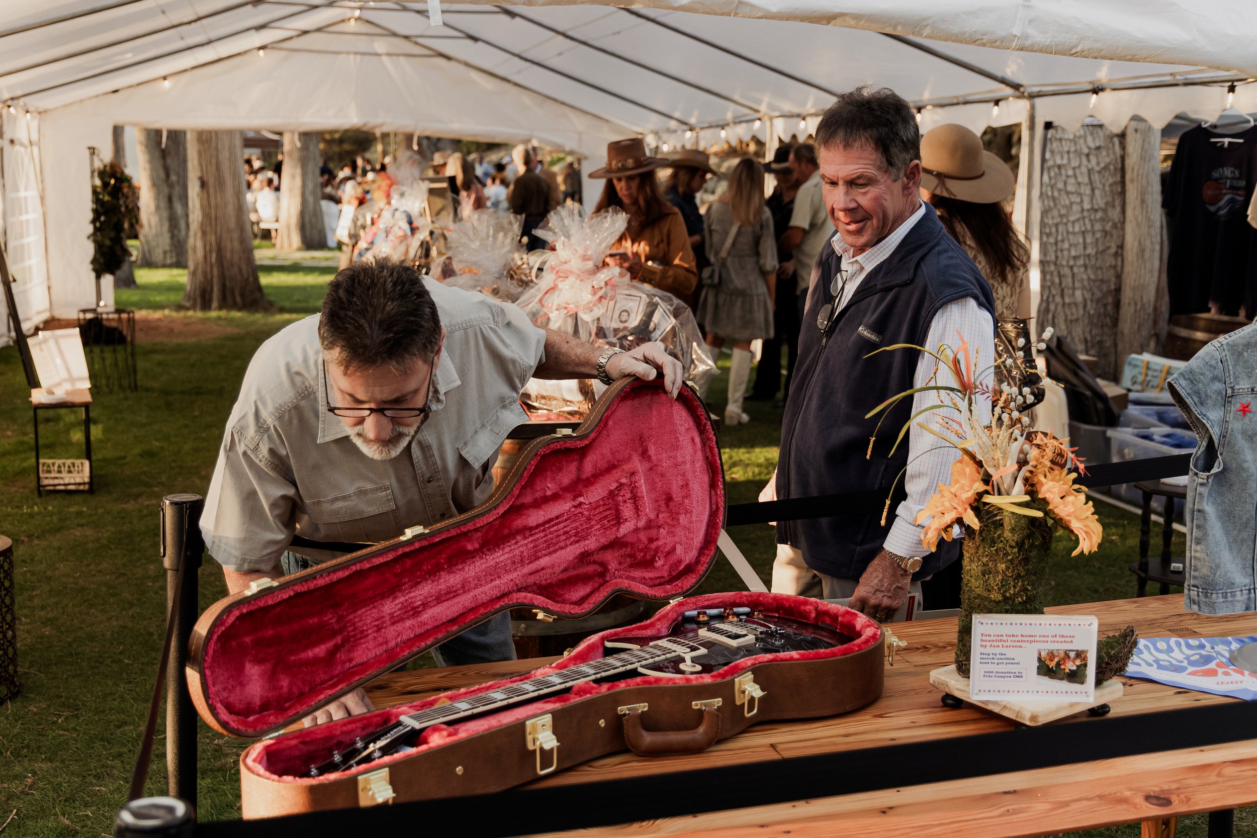 Two men examining a vintage guitar in a red velvet-lined case at an outdoor fair under a white tent, with other vendors and people in the background.