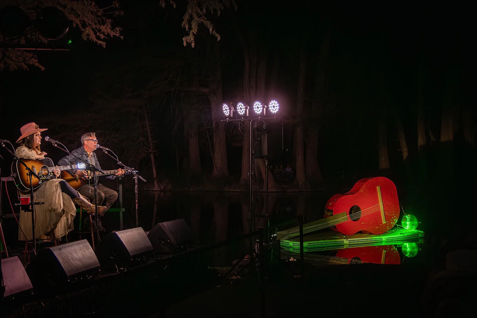 Two musicians playing acoustic guitars and singing on a stage outdoors at night, with stage lights, and a large, illuminated, red guitar sculpture lying on the ground next to the stage, reflected in a body of water.
