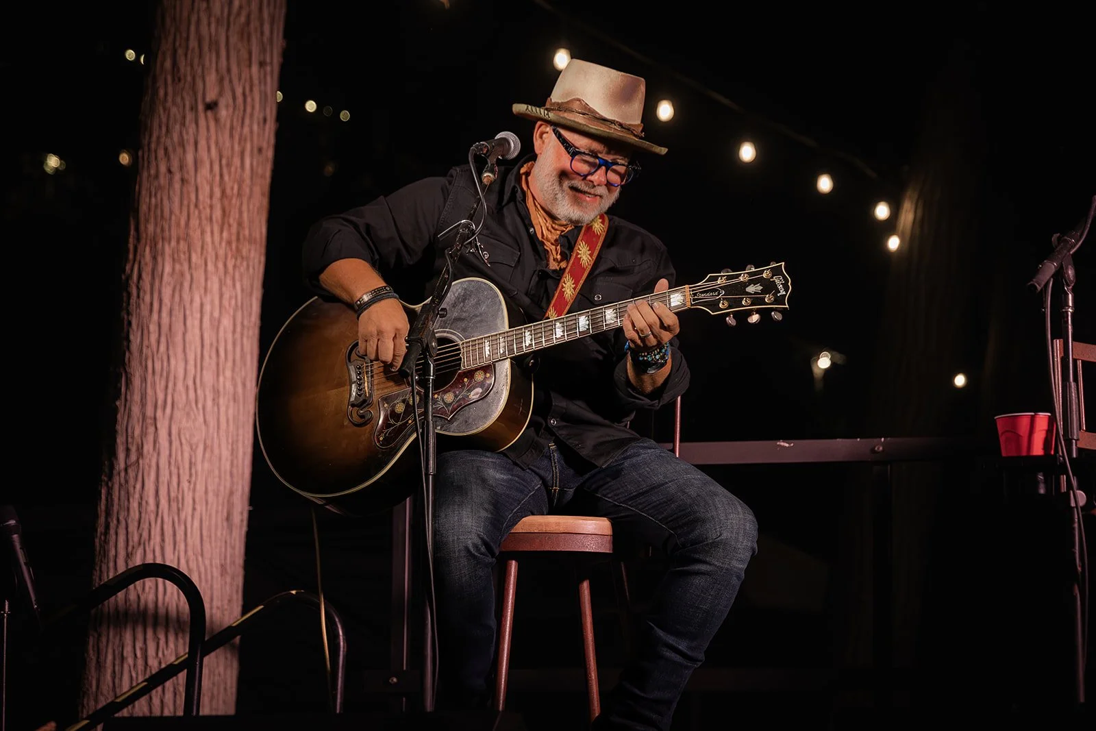 An older man with glasses, a hat, and a beard playing an acoustic guitar on stage at night, with string lights in the background.