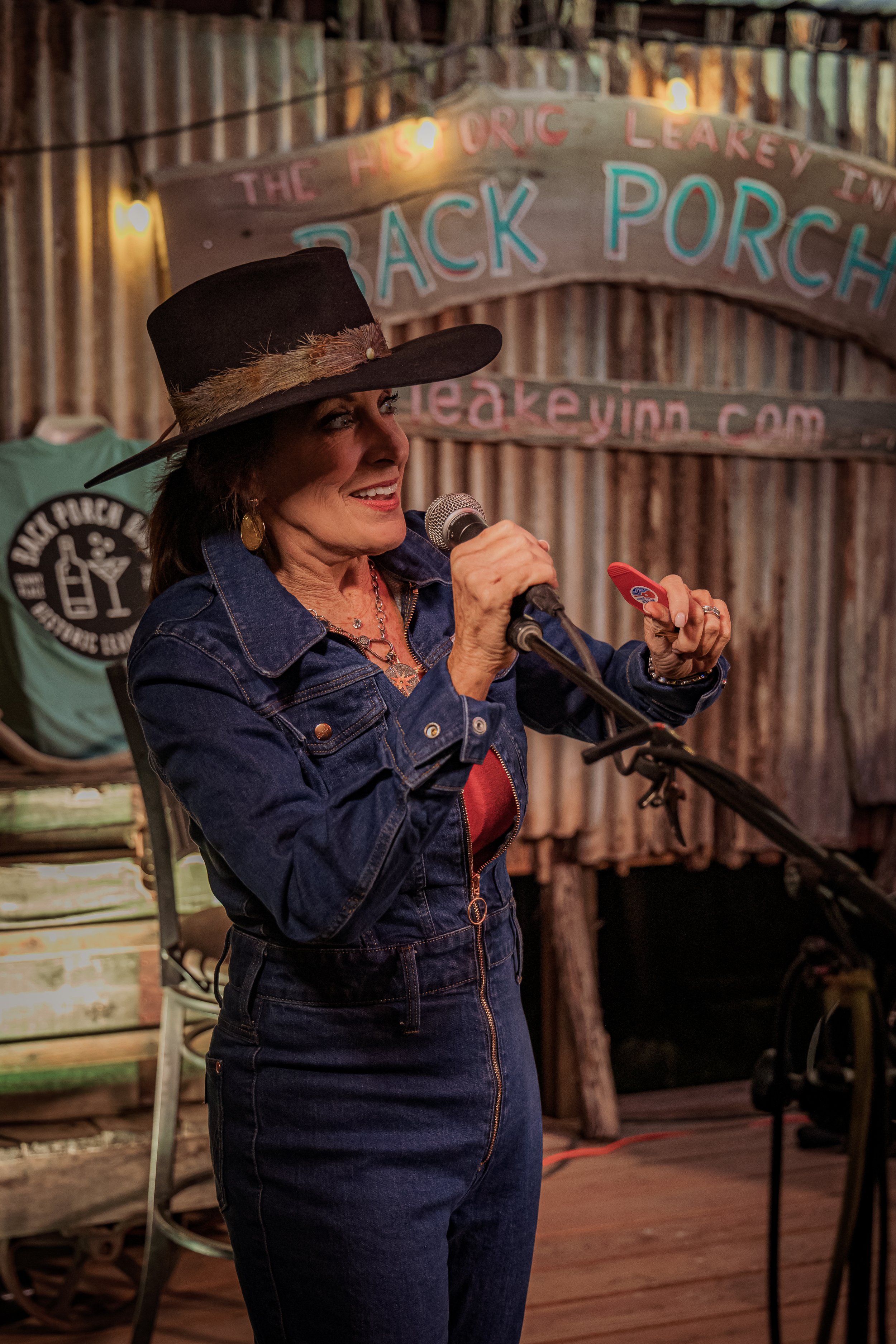 A woman wearing a black cowboy hat and denim overalls holding a microphone and speaking on stage at a rustic venue with wooden decor, in front of a sign that reads "The Hickory Leakey Inn" and "Back Porch".