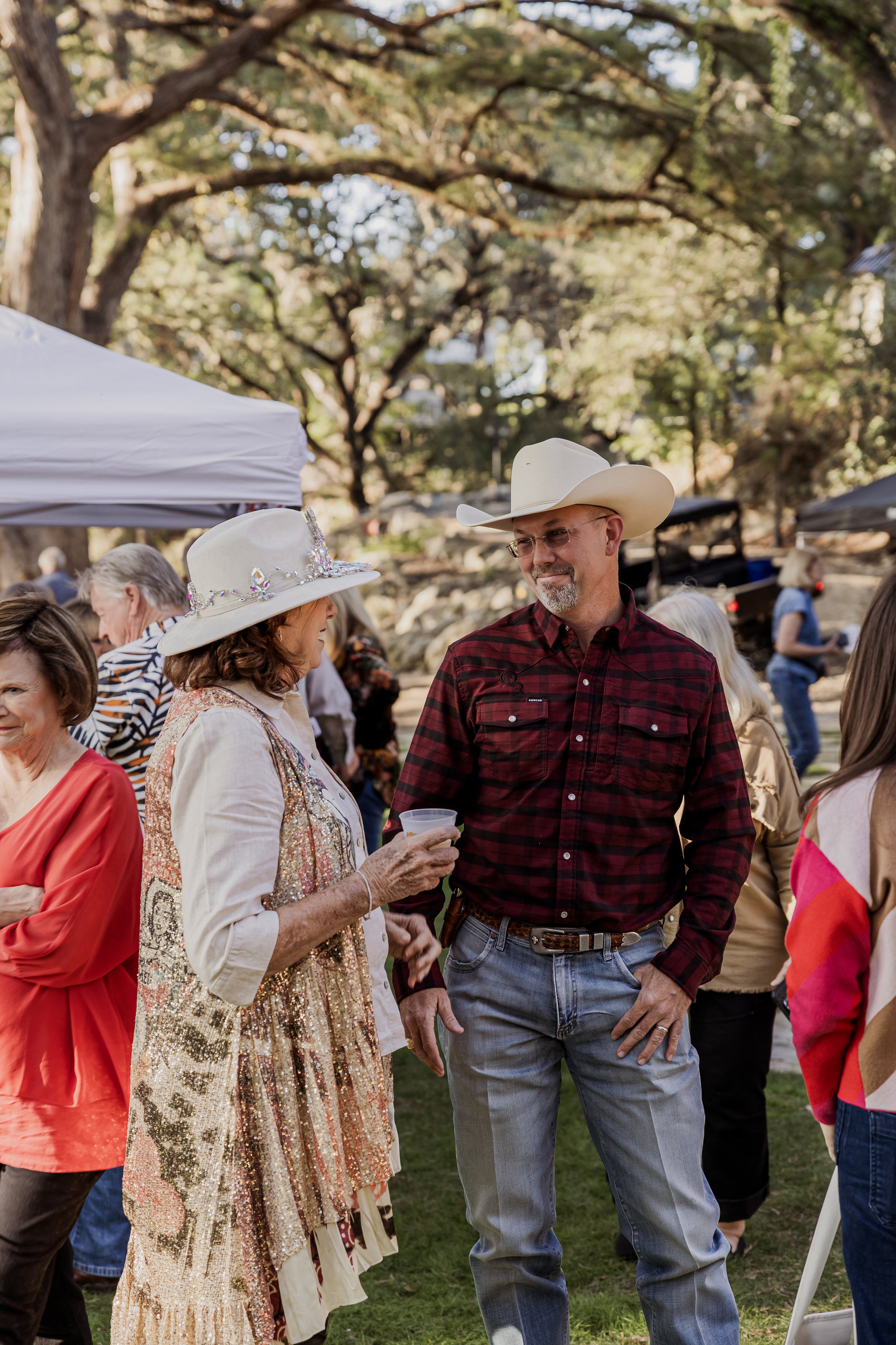 A woman and man wearing cowboy hats are talking at an outdoor gathering with trees in the background.