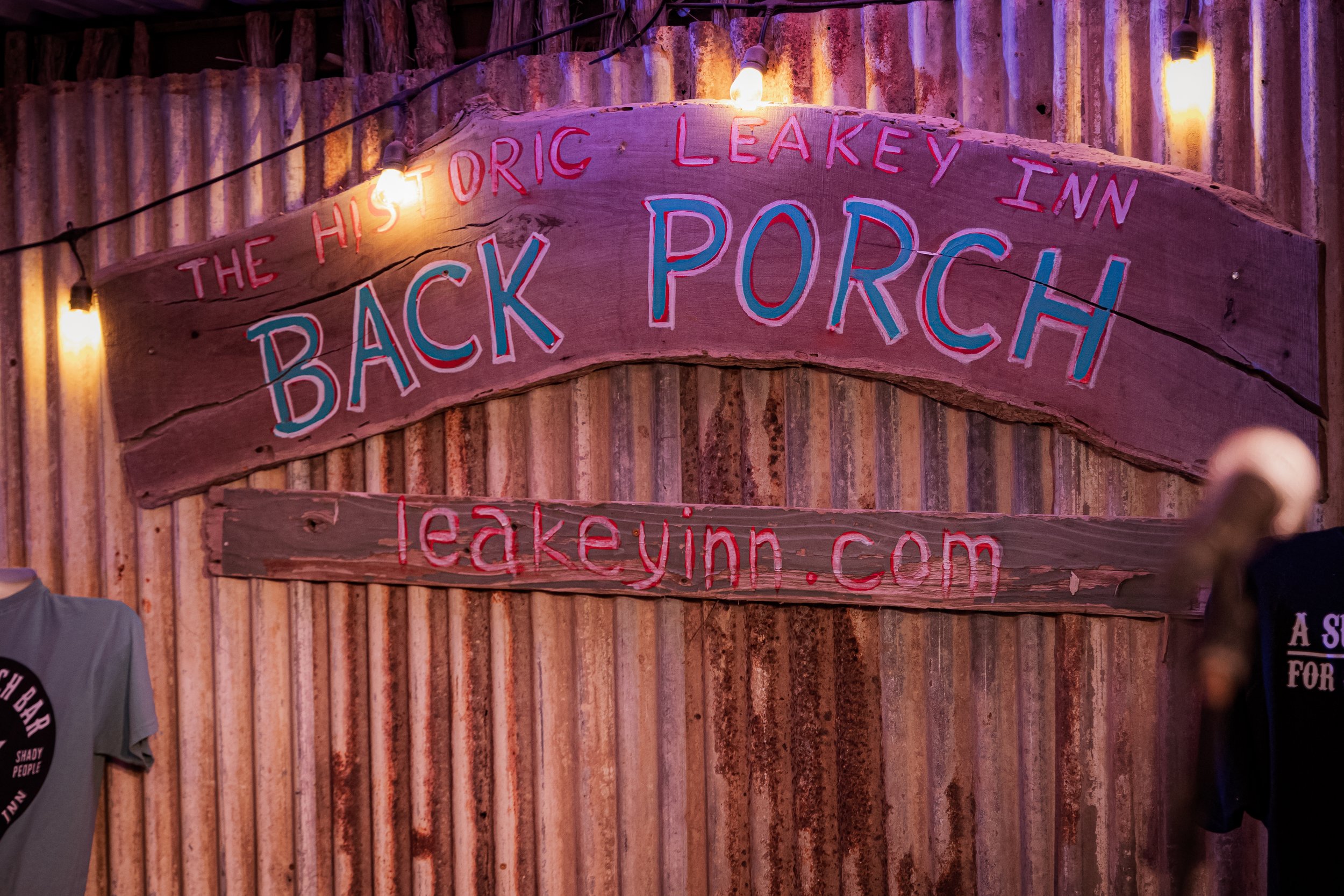 A wooden sign with colorful lettering that reads 'The Historic LeakeY Inn, Back Porch, leakeinn.com' on a rustic corrugated metal wall, illuminated by string lights.