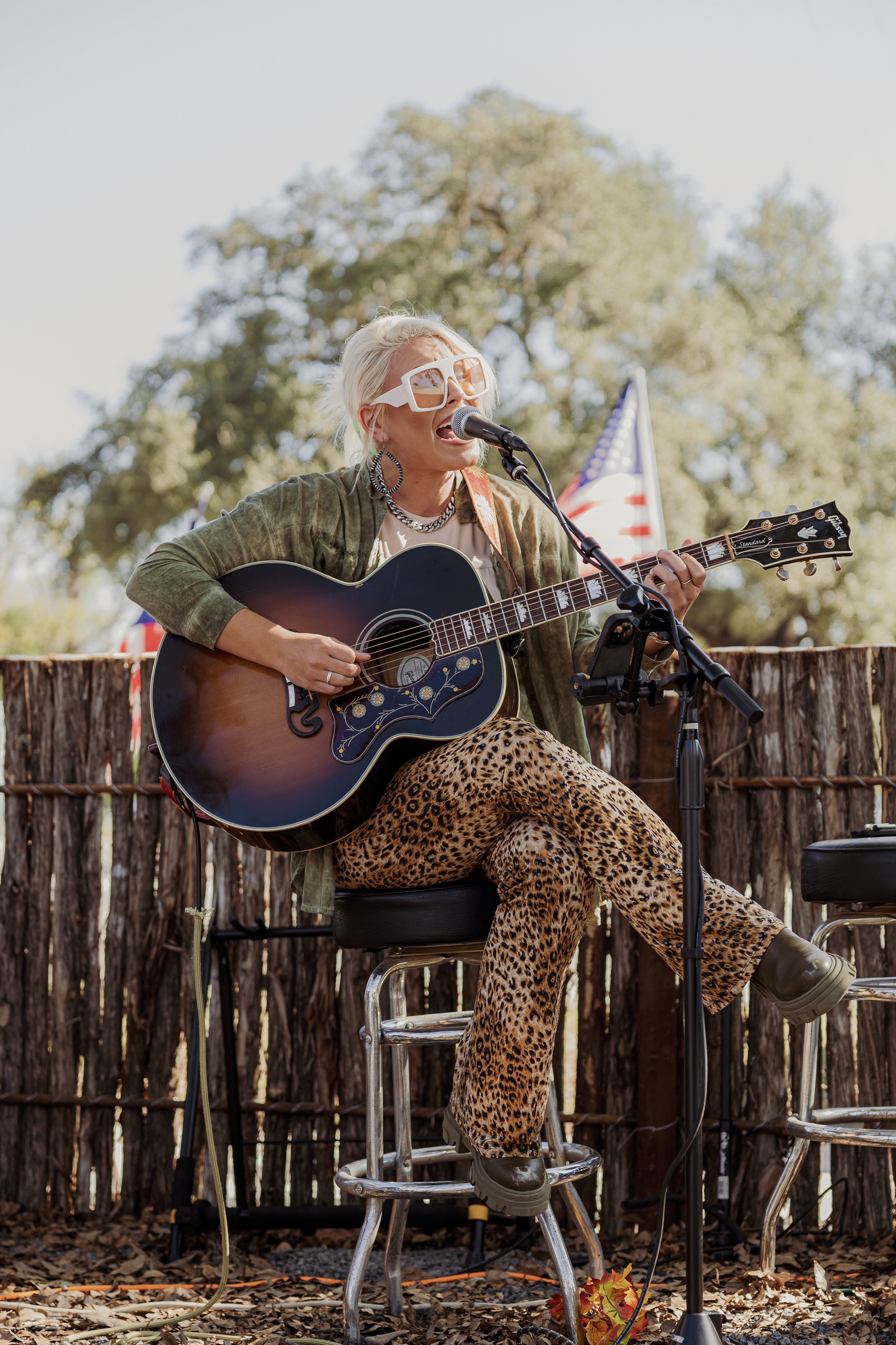 A woman with blonde hair, wearing oversized white sunglasses, patterned leopard print pants, a green jacket, and layered jewelry, is singing and playing an acoustic guitar during an outdoor performance with American flags in the background.