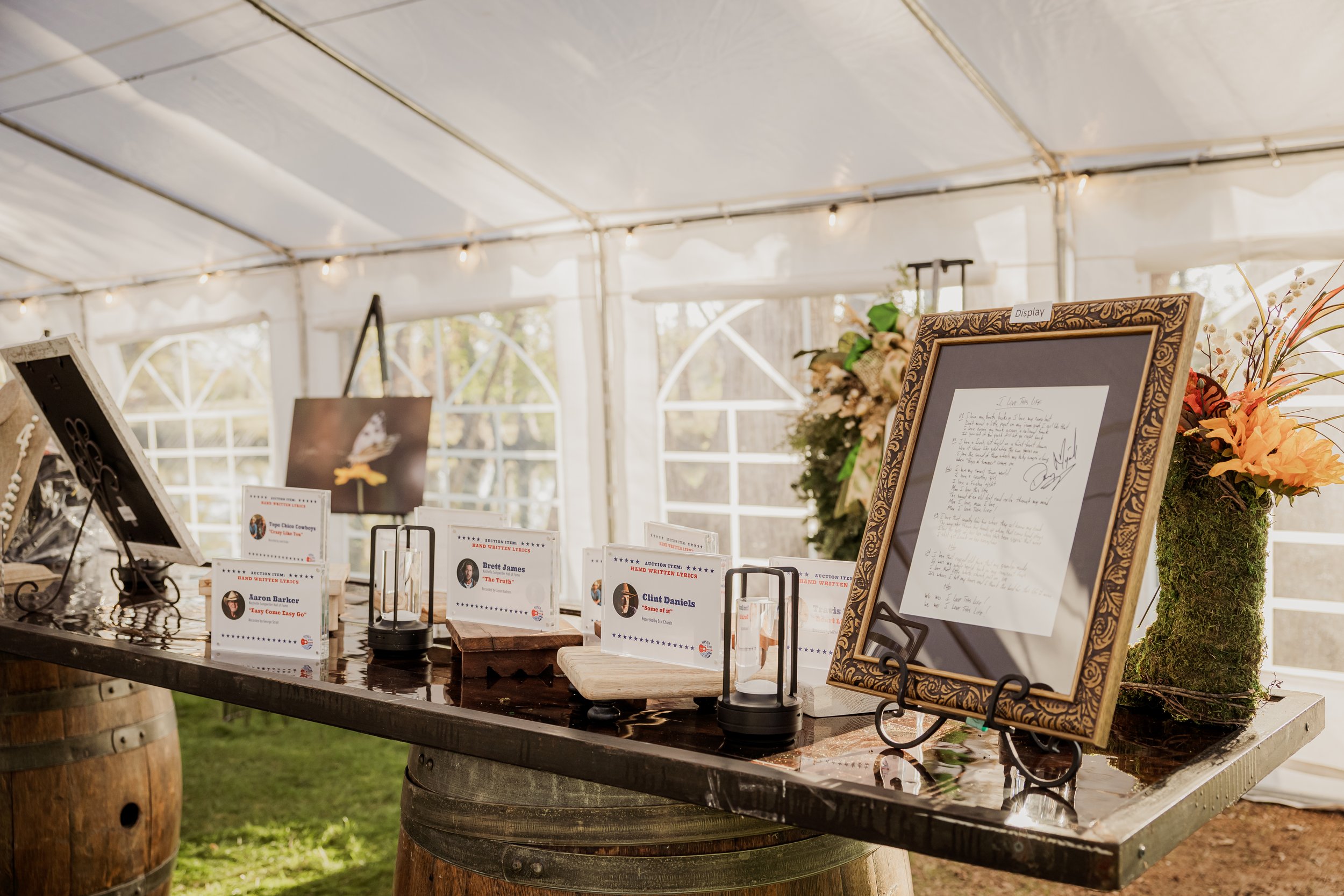 Display table with signed letters, framed artwork, flowers, and name cards in a tent setting for an event or auction.