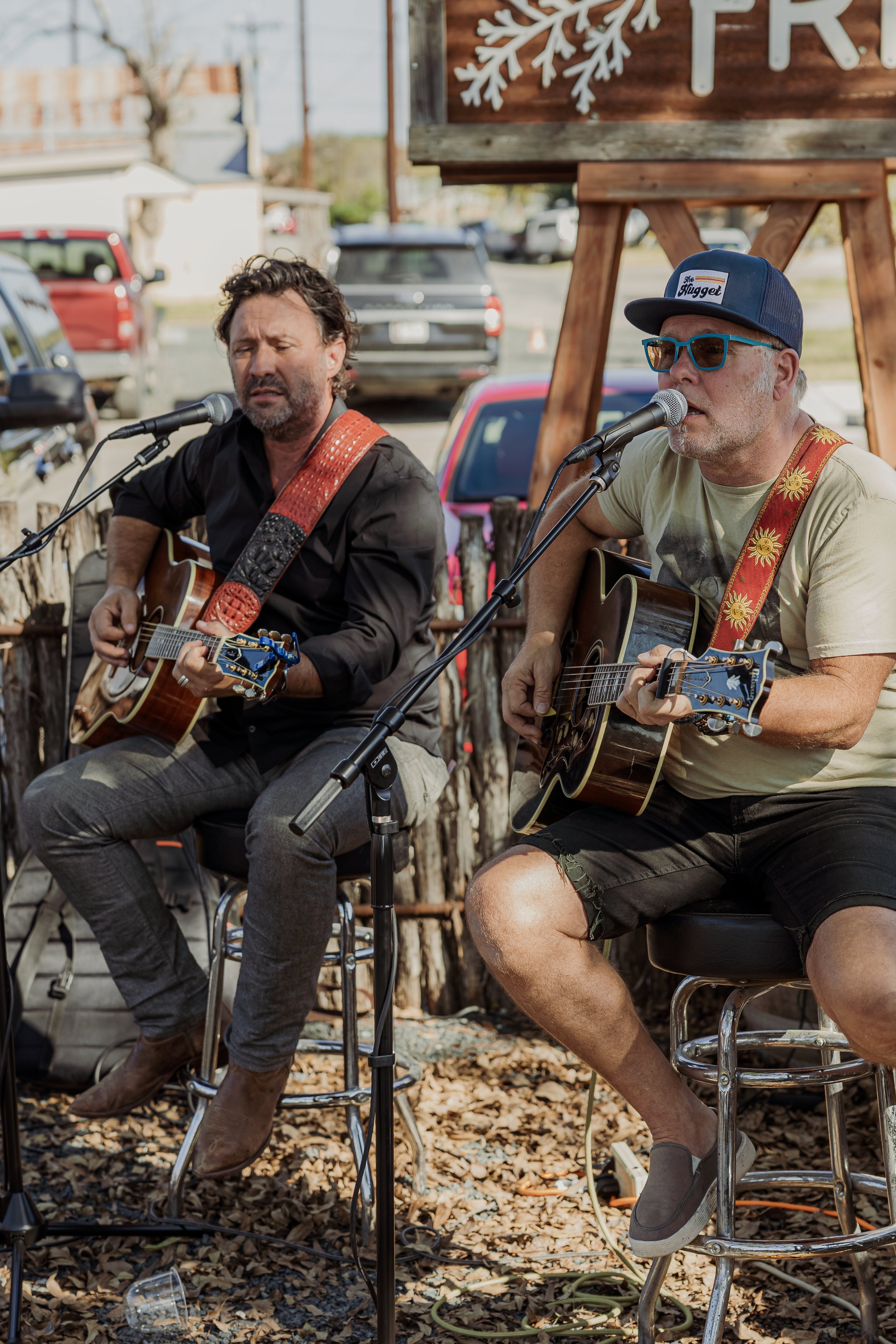Two men playing acoustic guitars and singing into microphones outdoors during the daytime.