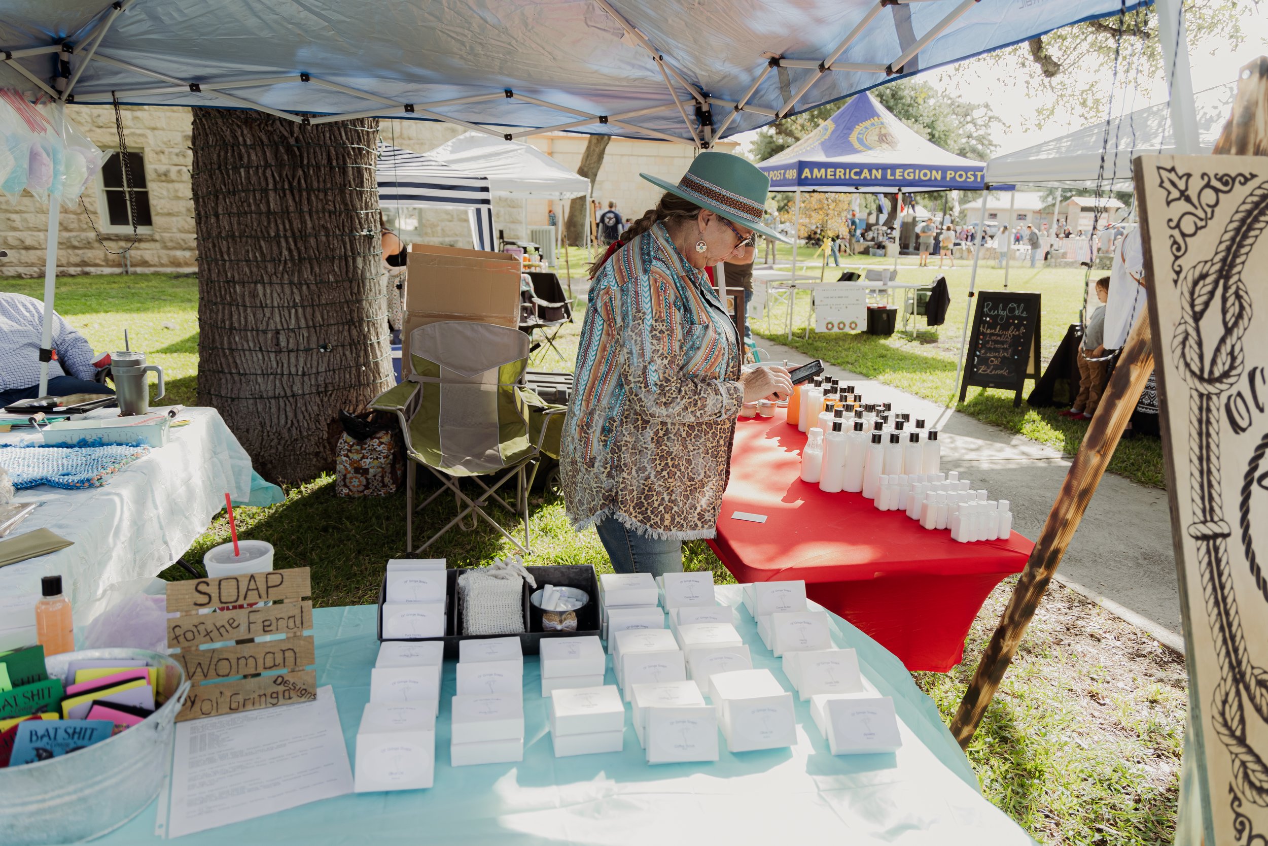 A woman at an outdoor market stall is arranging bottles on a red table, with soap bars on a blue table in front of her. She is wearing a teal hat, glasses, and a patterned blouse. Other market tents and people are visible in the background.