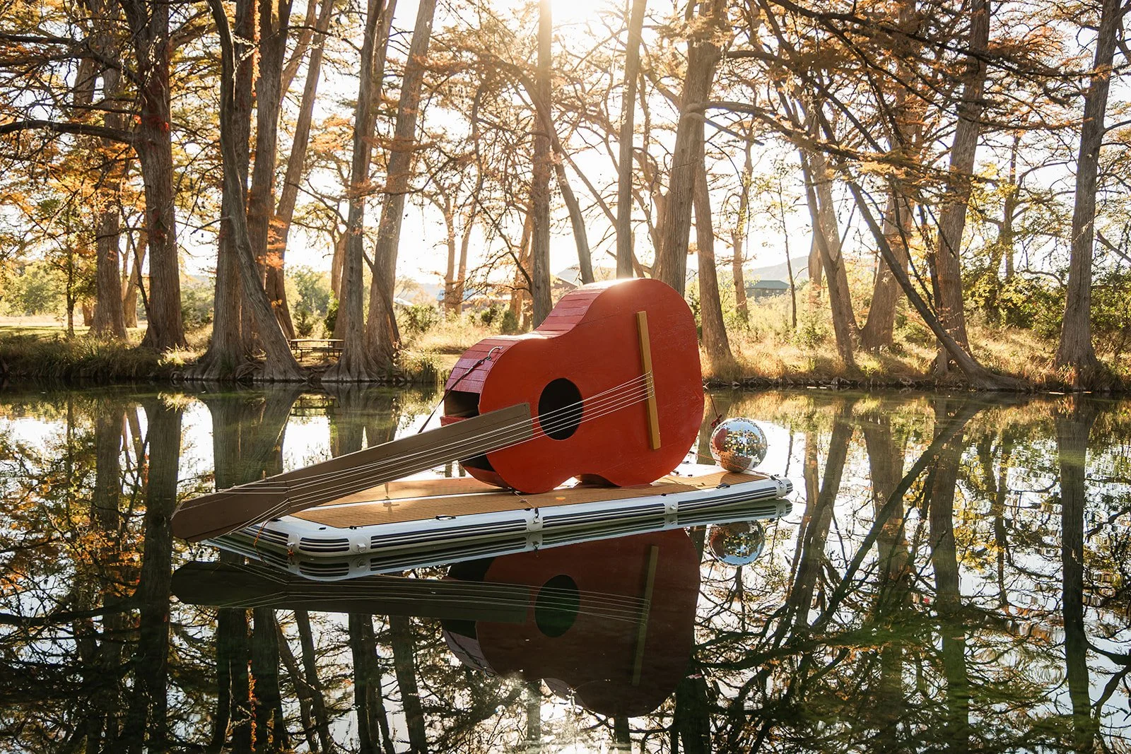 A large red guitar-shaped float on a floating platform in a pond, with a reflective surface and a forested background with trees and sunlight streaming through.