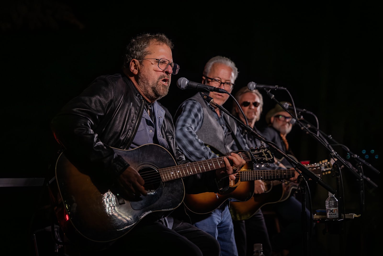 Four men seated, playing acoustic guitars and singing into microphones during a live performance at night.