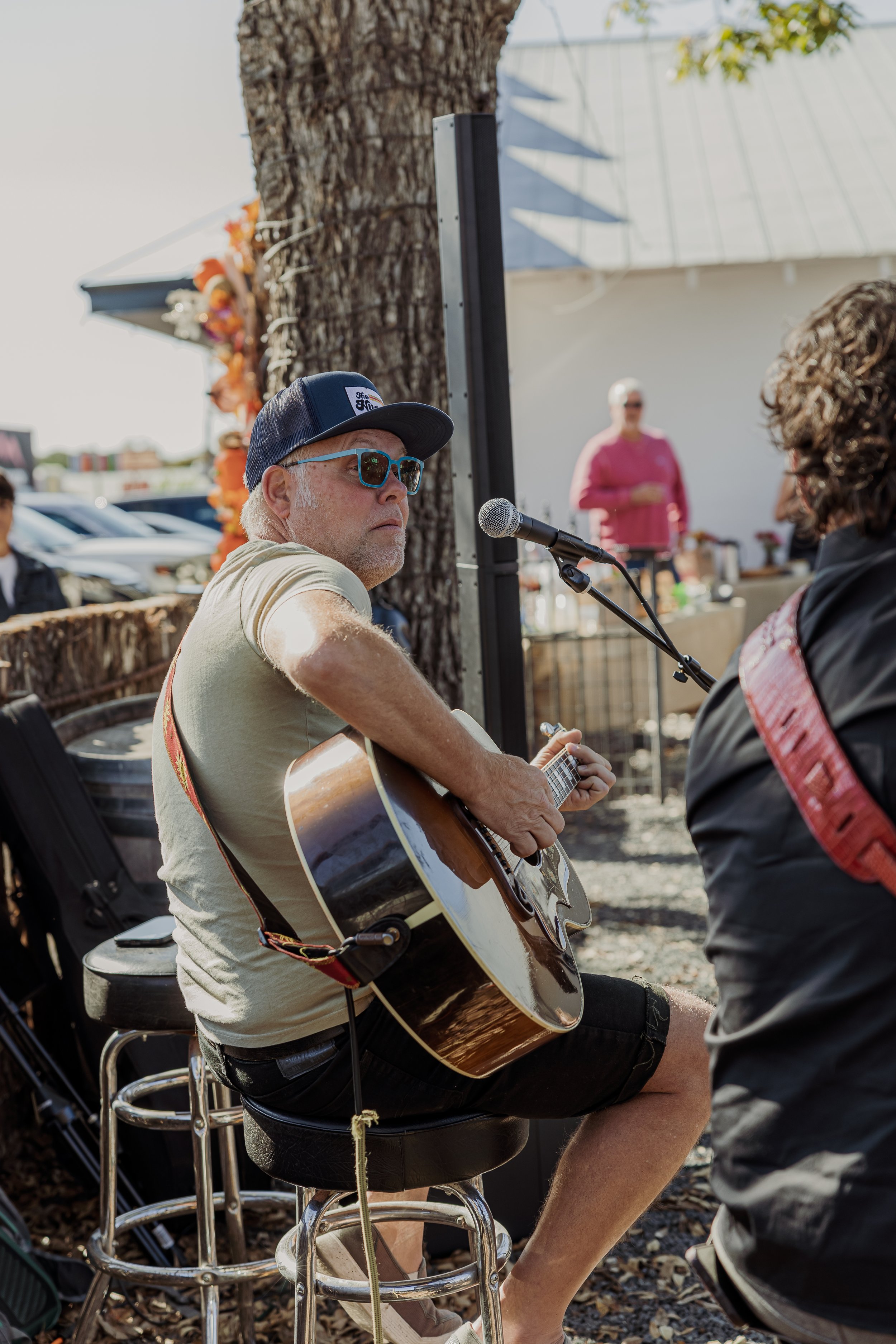 A man with sunglasses and a blue cap playing an acoustic guitar at an outdoor event.