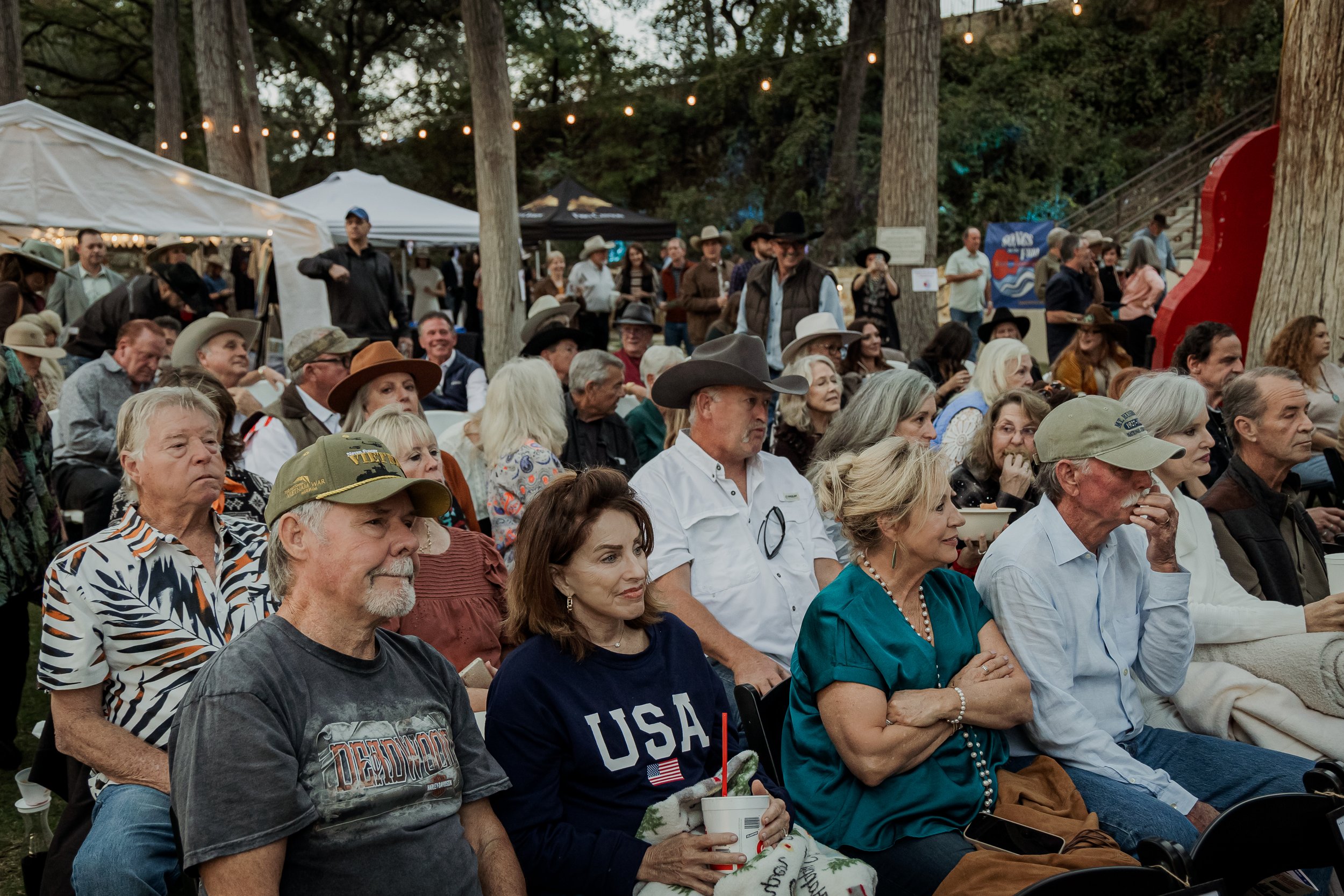 Audience seated outdoors at an event with trees, tents, and string lights overhead.