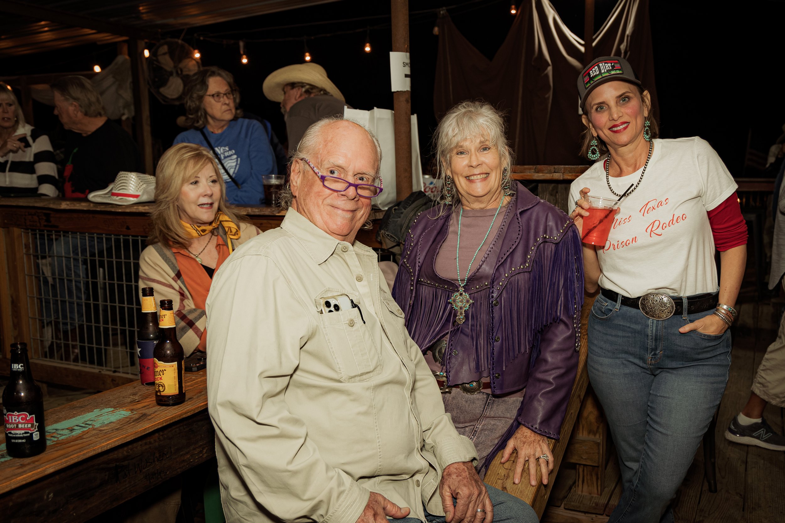 Group of people at a bar or restaurant, enjoying drinks and socializing, with a woman wearing a white "Miss Texas Prison Rodeo" T-shirt in the foreground.