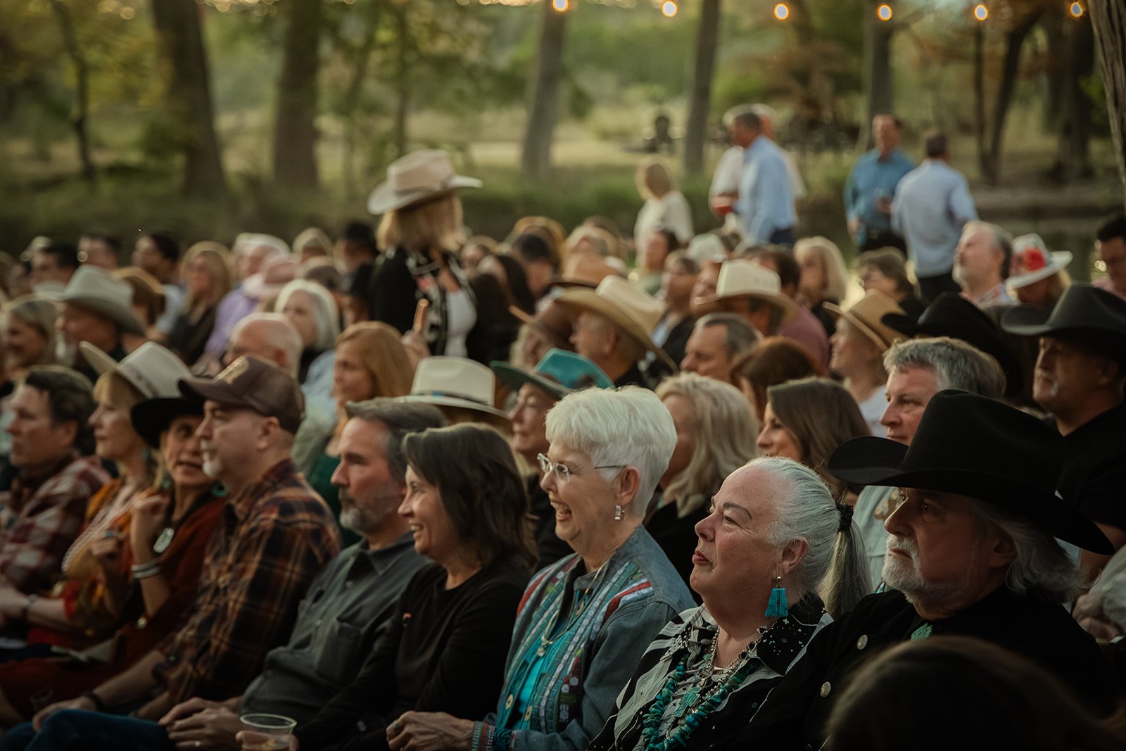 Crowd of people seated outdoors in a park, attending an event, many wearing cowboy hats and western-style clothing, with trees and string lights overhead.