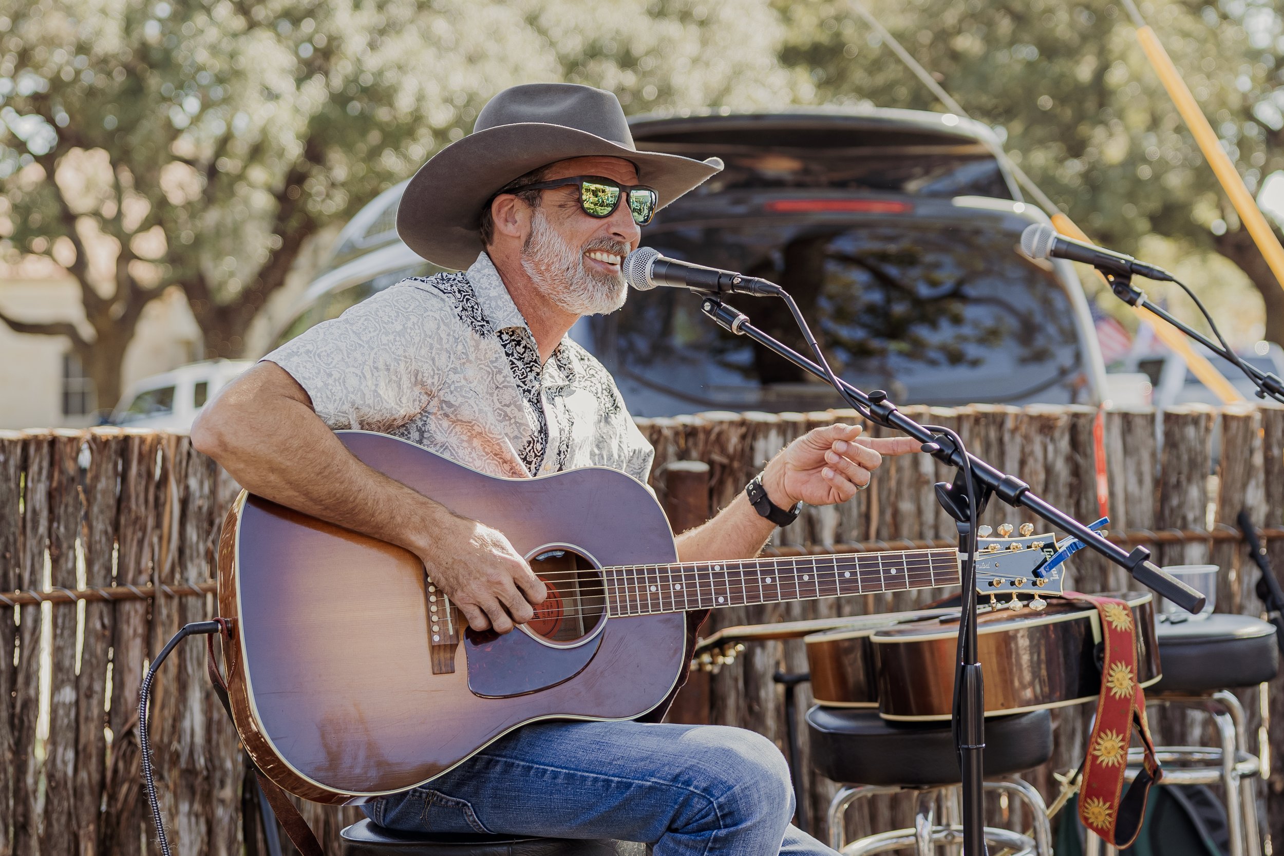 A man wearing a hat and sunglasses plays an acoustic guitar and sings into a microphone at an outdoor event. He is smiling and seated in front of a rustic wooden fence, with a black van and trees in the background.