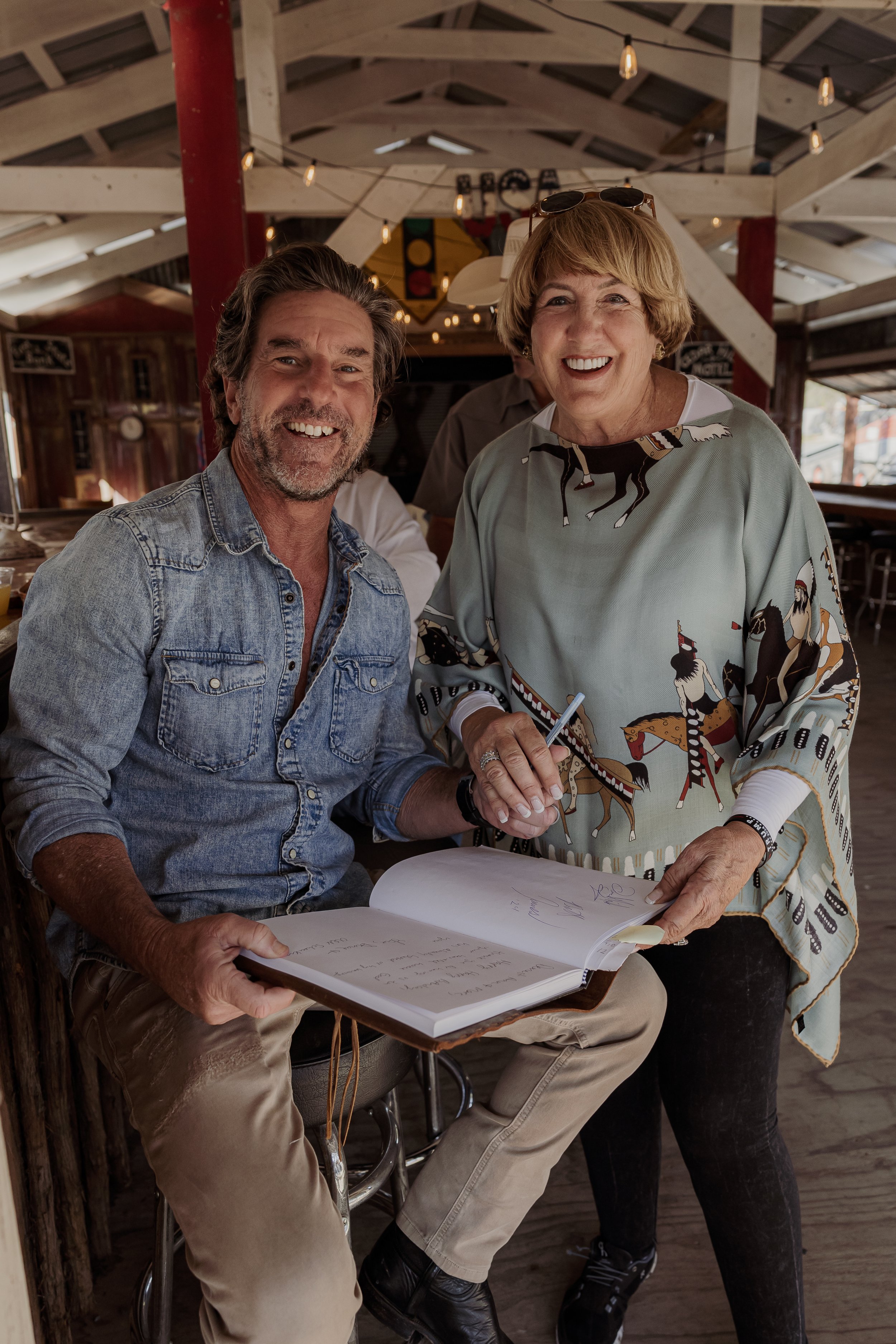 A man sitting on a barstool signing a guest book with a woman standing next to him, both smiling. The man has gray hair and a beard, wearing a denim shirt. The woman has short blonde hair and is wearing a colorful, patterned top. They are in a rustic