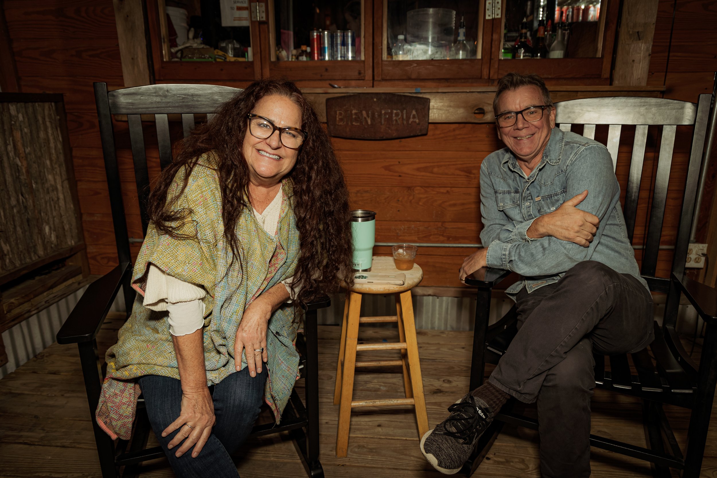 A smiling woman with long curly hair and glasses sitting on a black chair, and a smiling man with glasses and short hair sitting on another black chair, in a wooden rustic room with a small wooden table between them holding a travel mug and a glass o