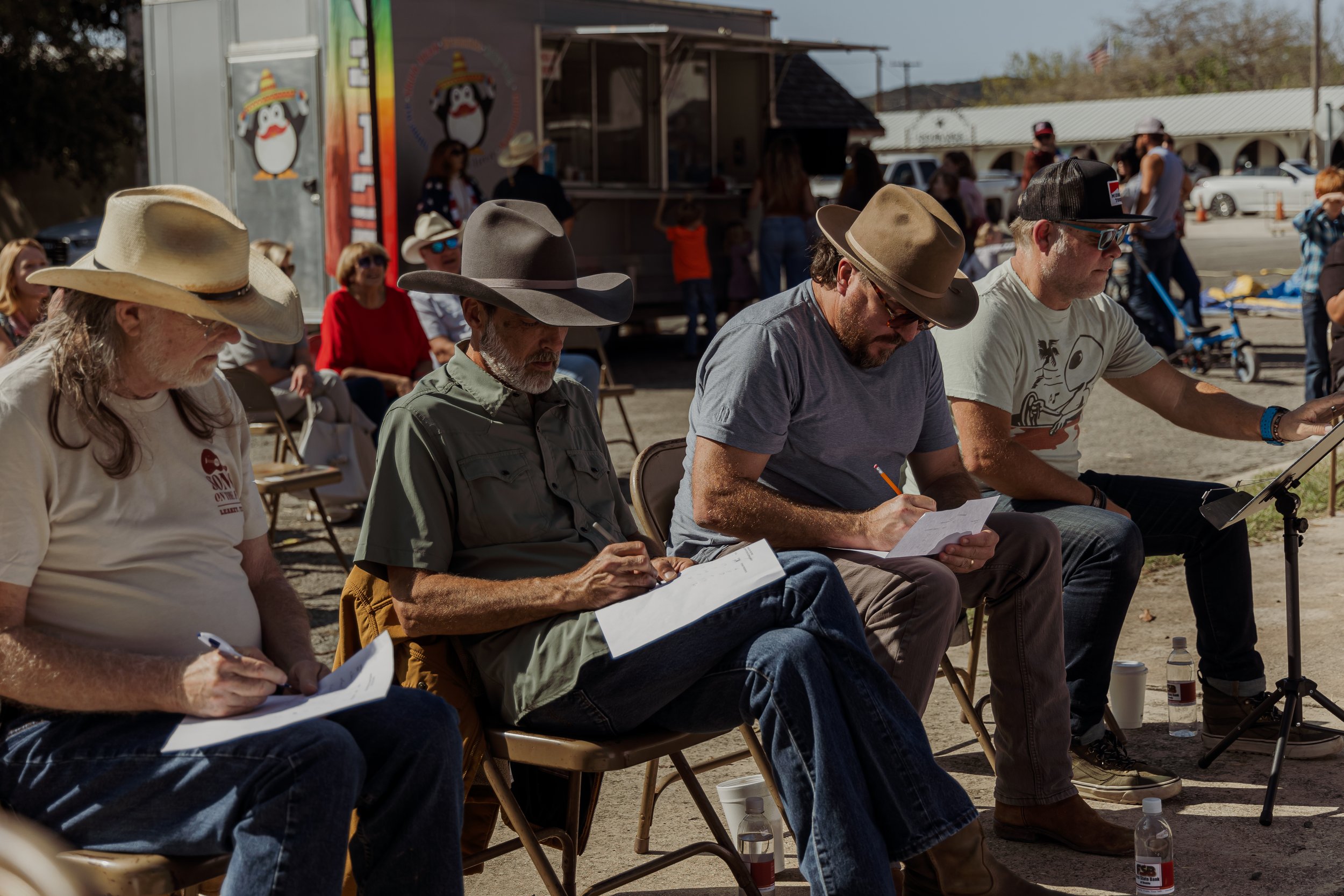 Four men sitting outdoors in a row, writing or taking notes on paper, with an audience and a food truck in the background.