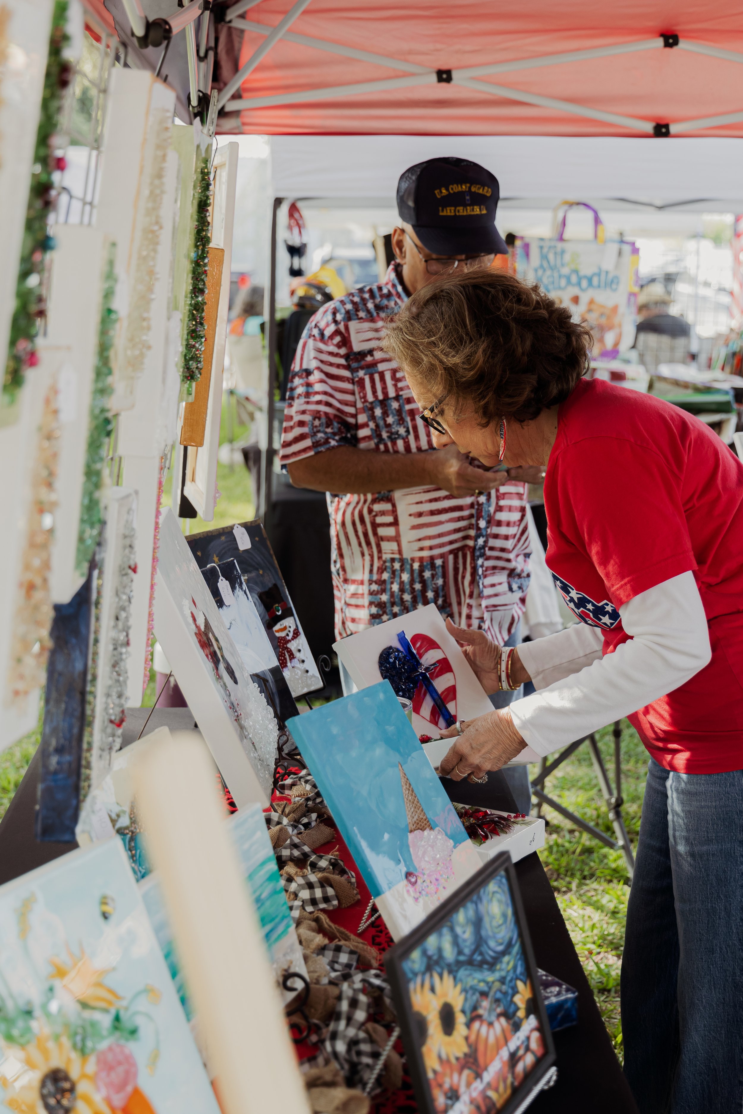 Two women at an outdoor craft market, looking at artwork and jewelry on display under a red canopy.