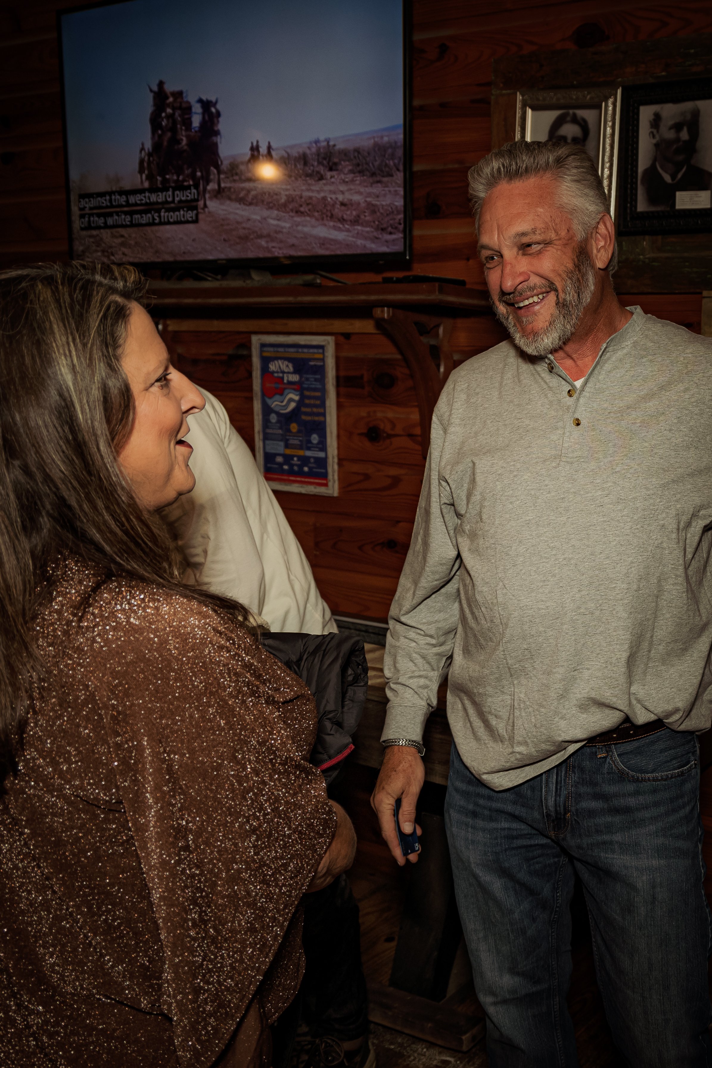 A woman and a man smiling and talking inside a wooden-paneled room, with a television screen showing a Western scene and framed photos on the wall behind them.
