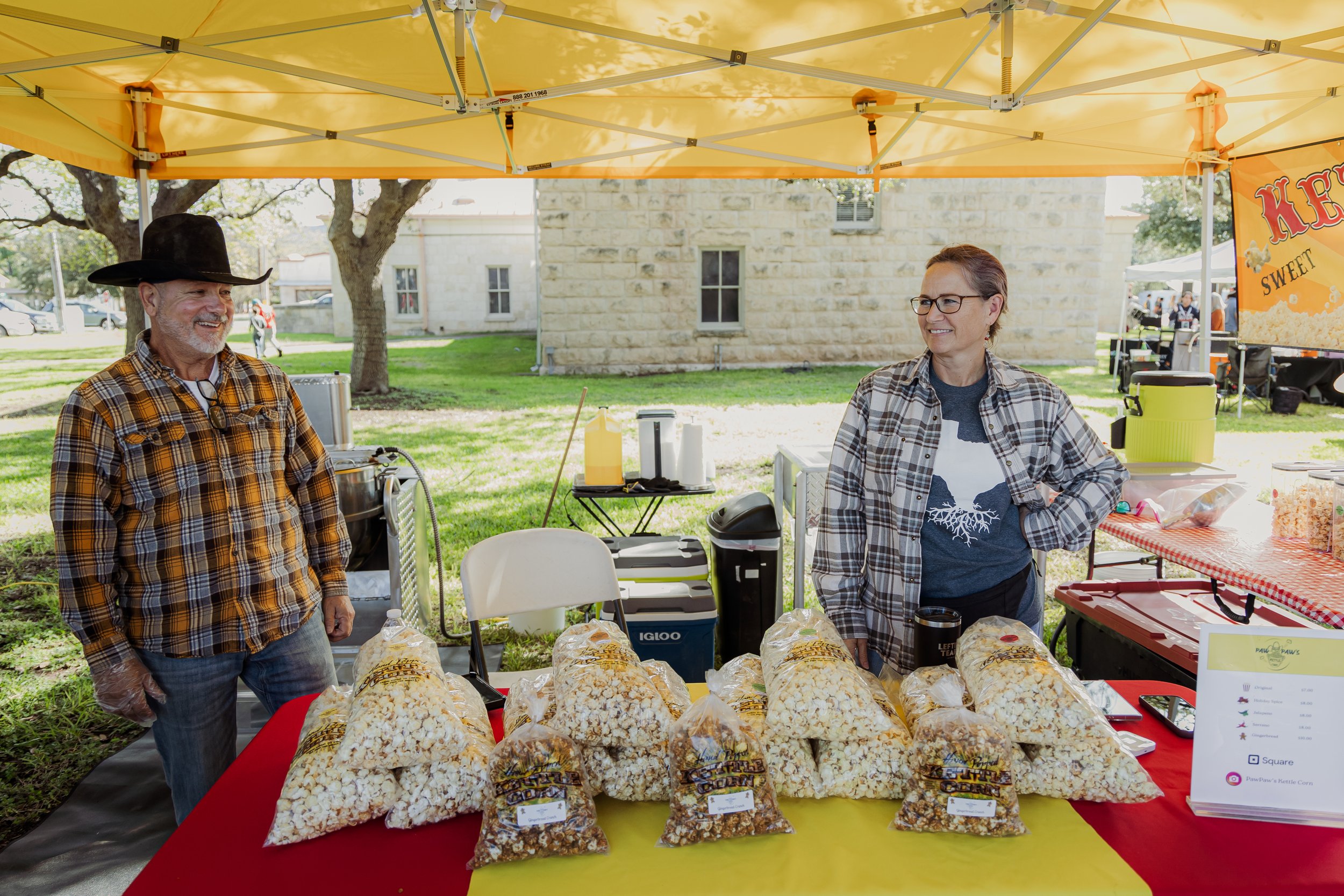 Two people at a popcorn stand, a man wearing a cowboy hat and plaid shirt, and a woman wearing glasses and plaid shirt, surrounded by bags of popcorn on a red and yellow table at an outdoor fair.