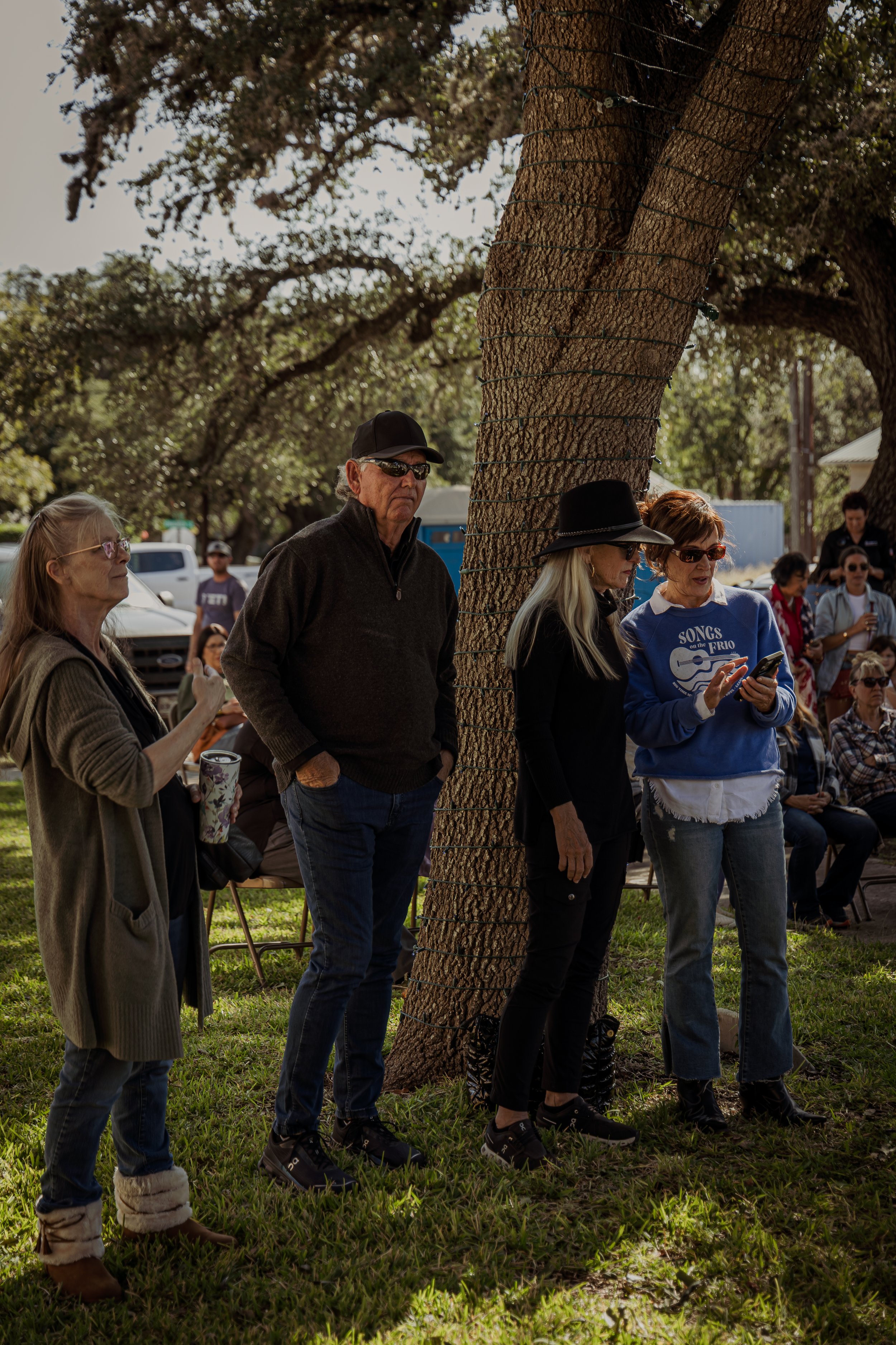 Group of people standing under a large tree in a park, some looking at their phones and others observing or listening, with additional people seated and standing in the background.