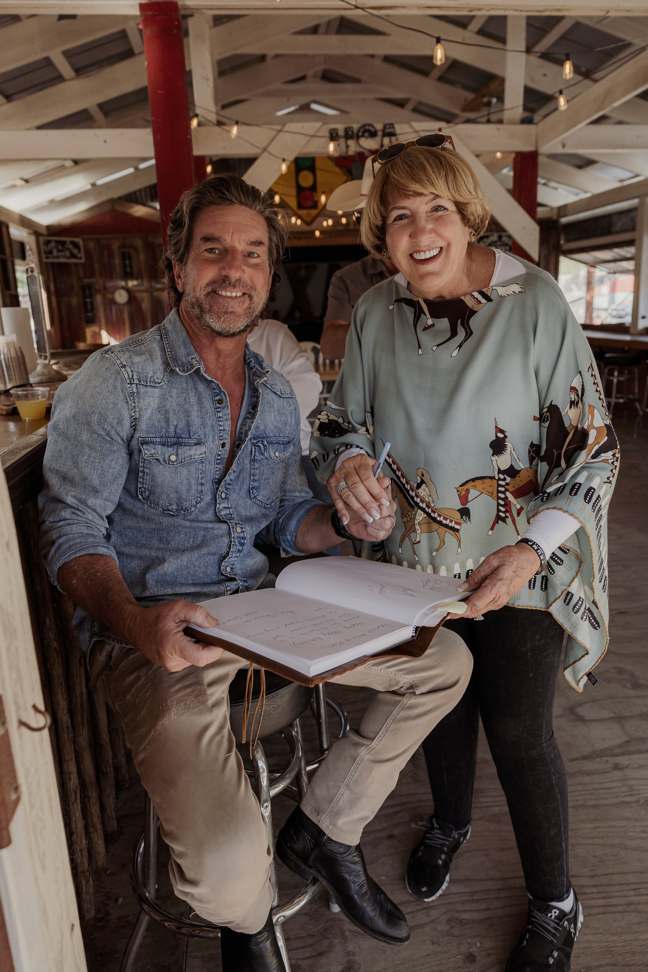 A man seated at a bar holding a large open guestbook and a woman signing it, both smiling in a rustic, sunlit restaurant with string lights overhead.