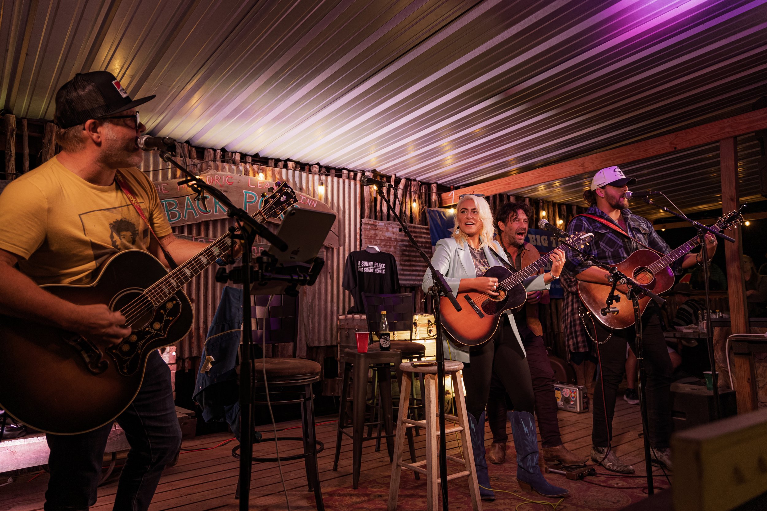 Four musicians performing with guitars on a stage decorated with string lights and a sign that reads "The Back Porch Inn."